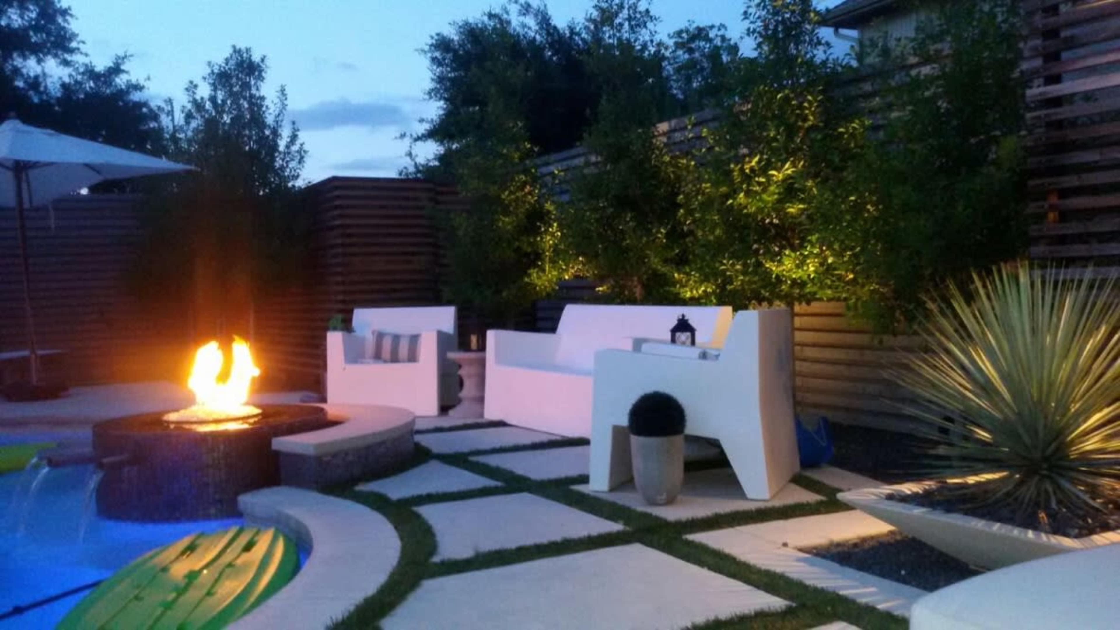 The scene features a cozy outdoor seating area beside a glowing fire pit, surrounded by greenery and soft lighting.