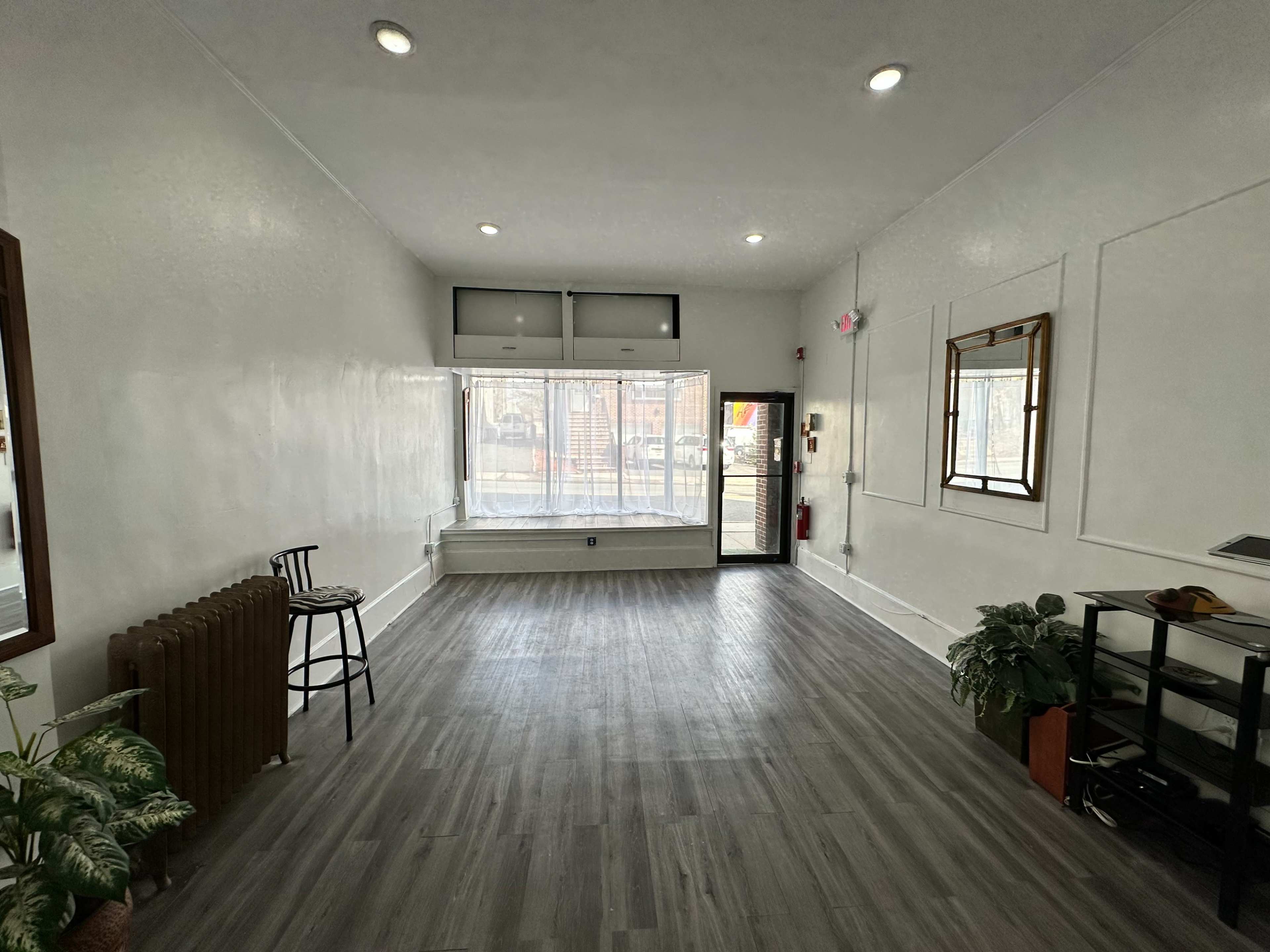The image shows a spacious, well-lit room with wooden flooring, a large front window, and minimal furnishings including a chair and a small table.