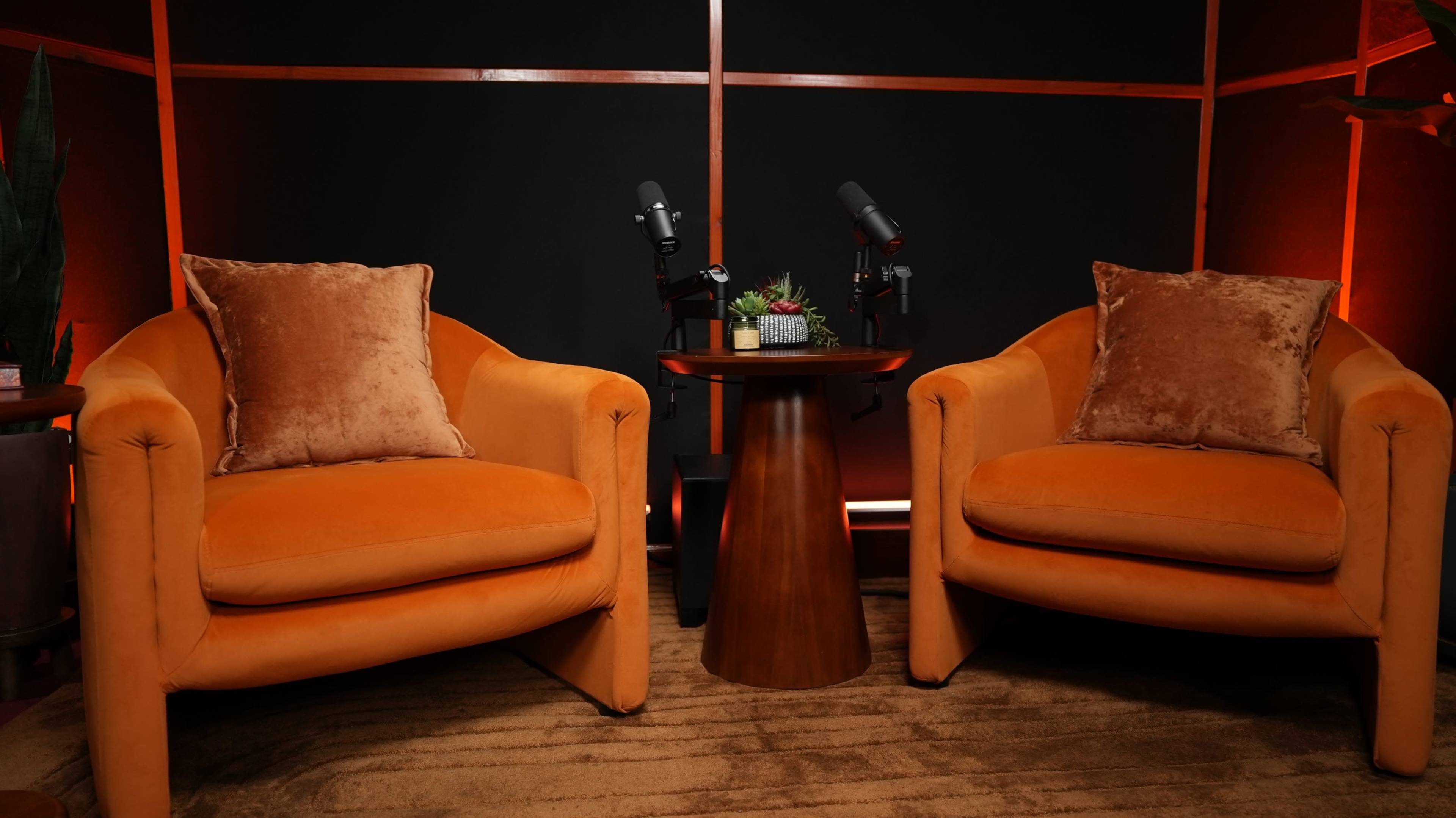 The image shows two orange sofas with plush pillows facing a wooden table, accompanied by microphones on the table against a dark backdrop.