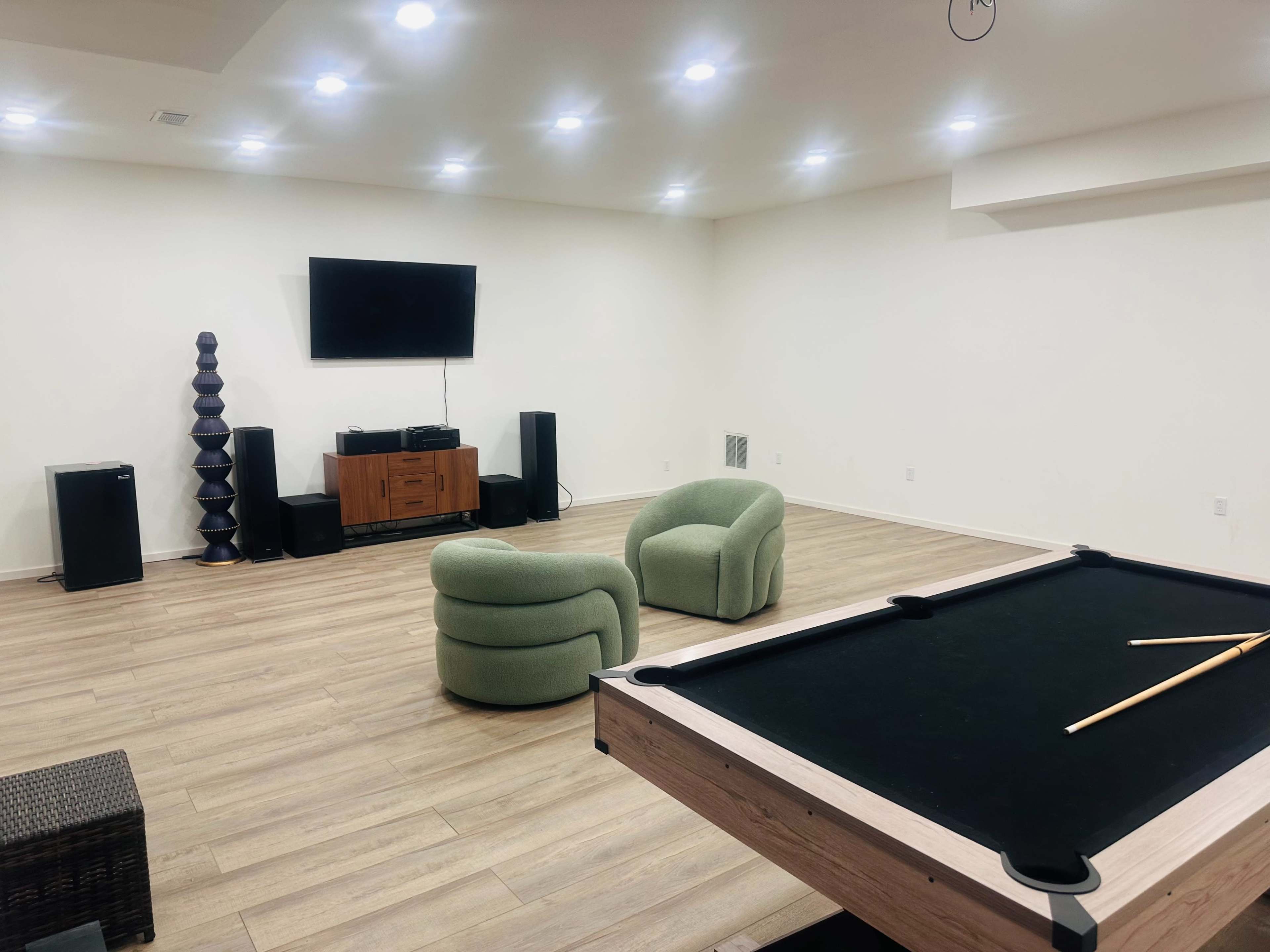 The image shows a spacious room with light wood flooring, featuring a pool table, two green armchairs, a television mounted on the wall, and audio equipment.