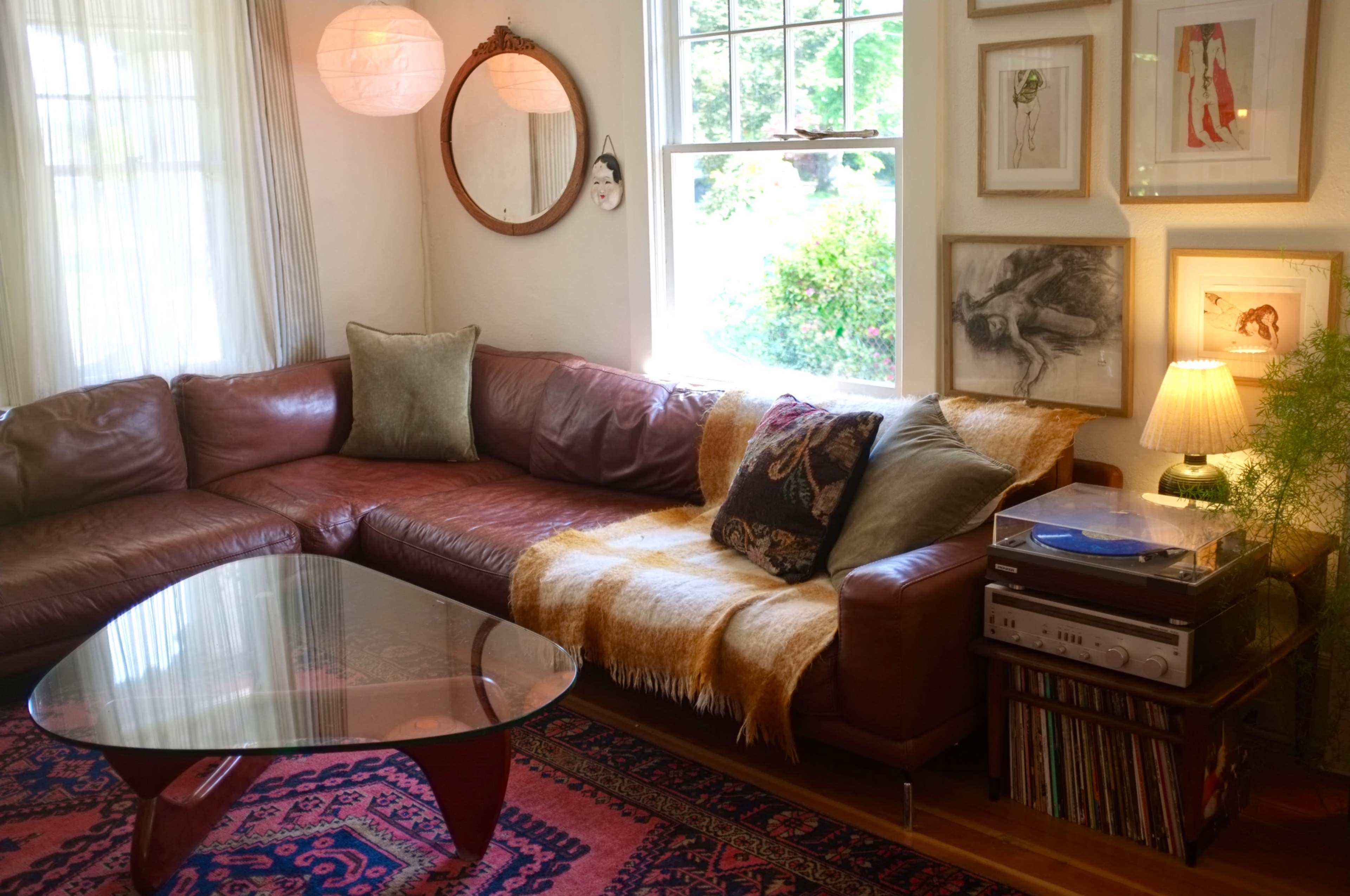 A cozy living room features a brown leather sectional couch with a soft throw, a glass coffee table, a record player next to a stack of vinyl records, and framed artwork on the walls.