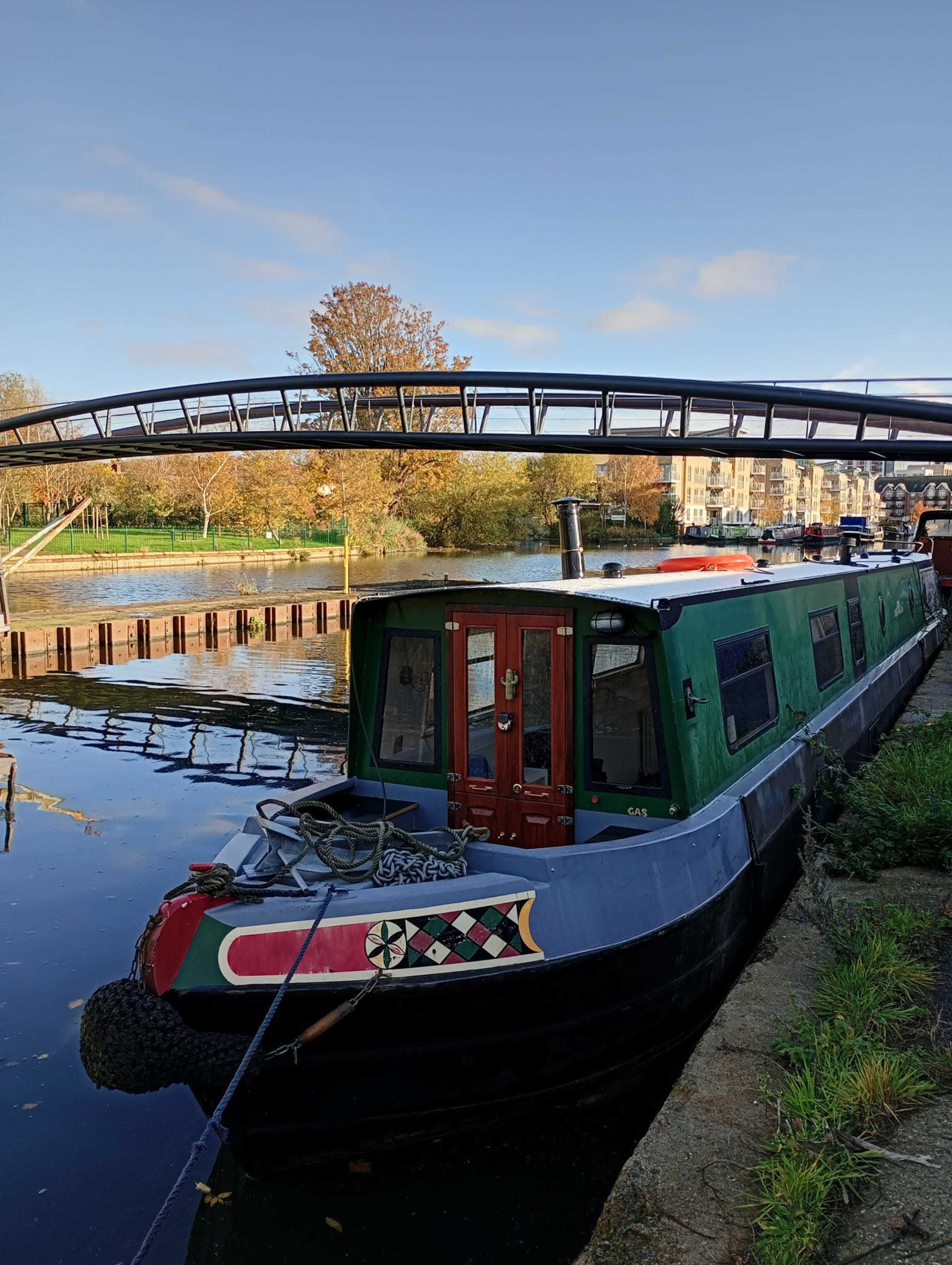 A narrowboat is moored alongside a canal beneath a curved footbridge, with trees and buildings in the background.