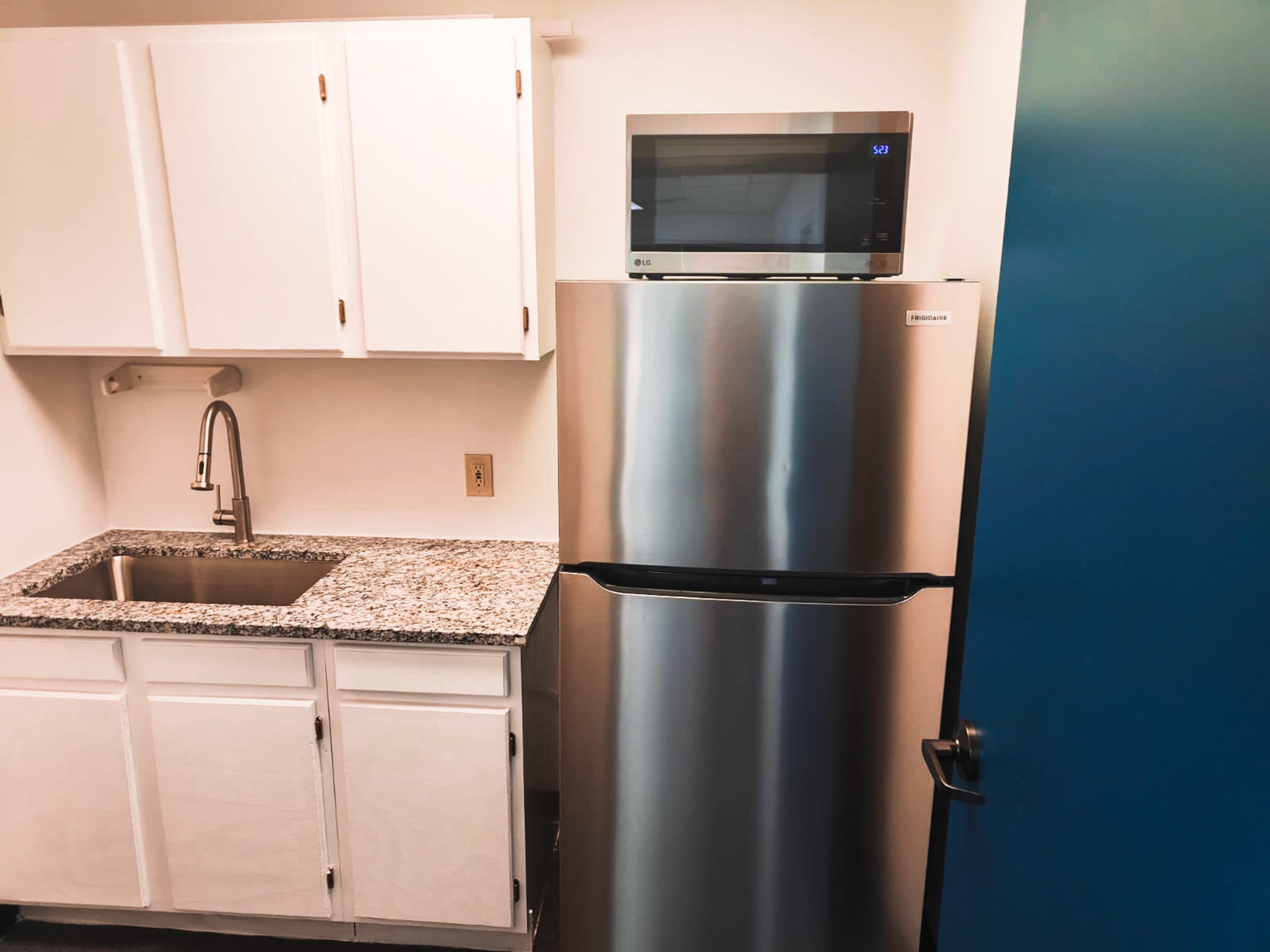 A kitchen area with a stainless steel refrigerator, a small microwave on top, a granite countertop, and white cabinets.
