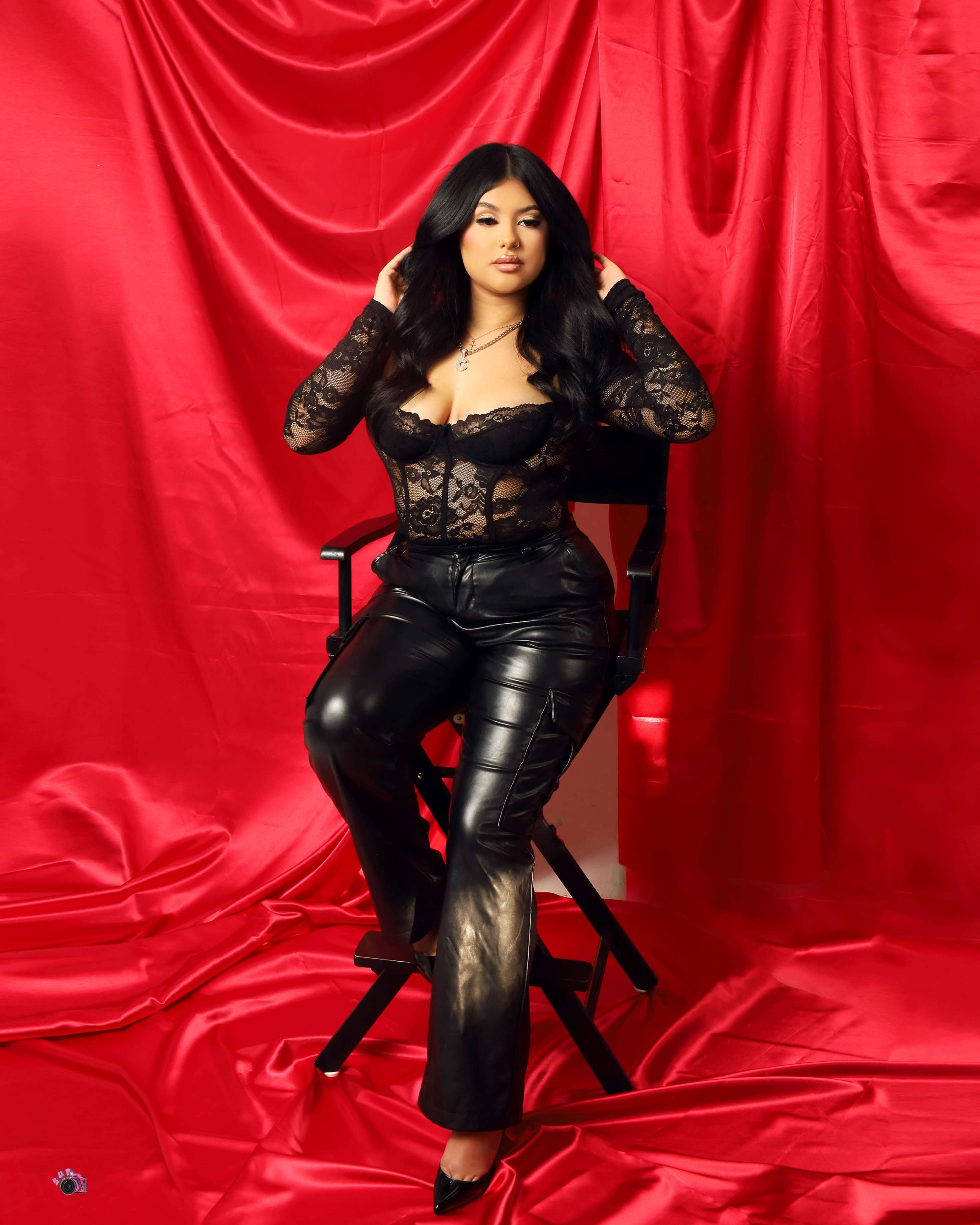A woman poses on a chair against a red backdrop, wearing a black lace top and shiny black pants.
