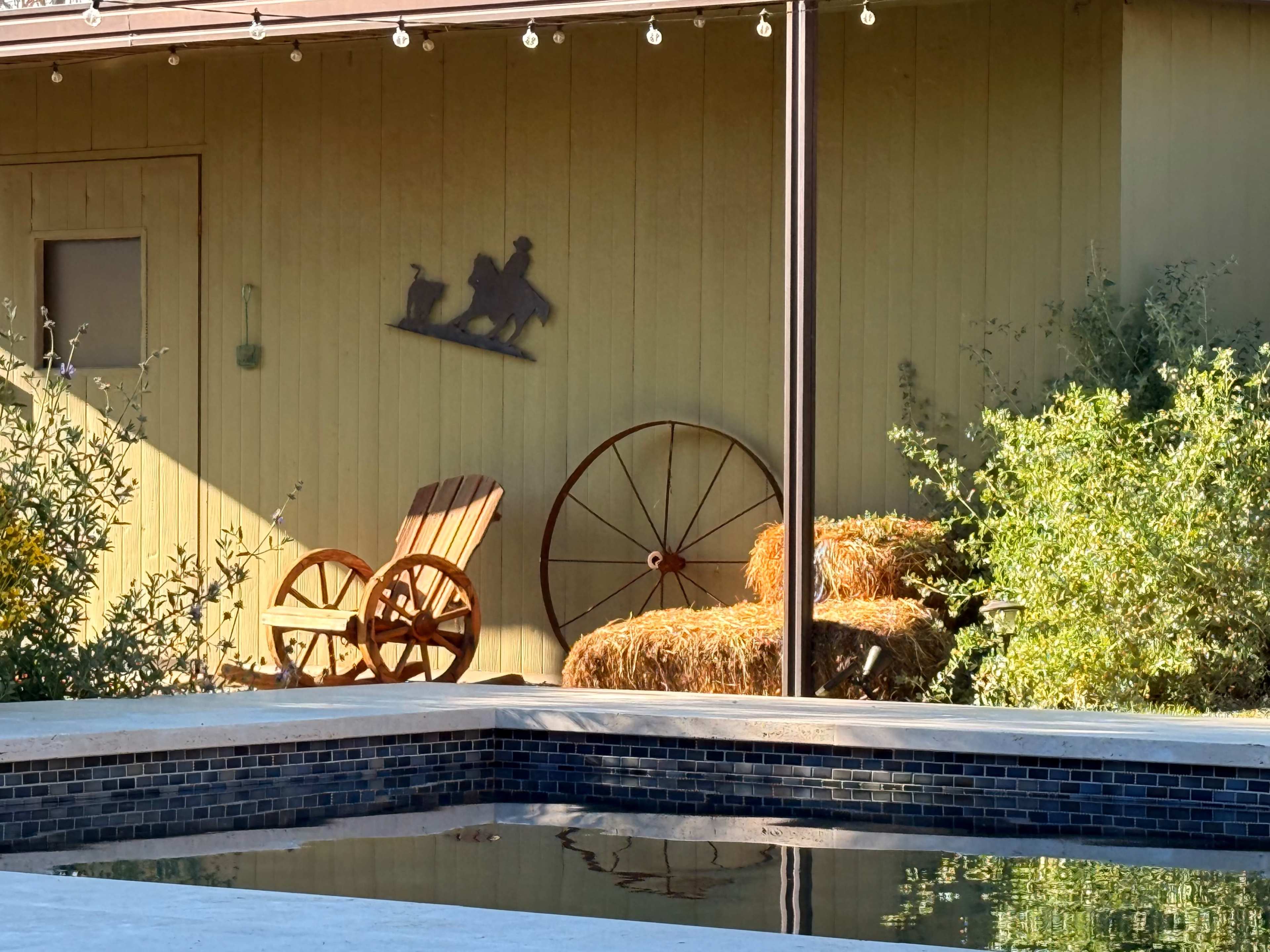 A wooden chair and a hay bale are placed near a pool in front of a yellow building with a decorative silhouette hanging on the wall.