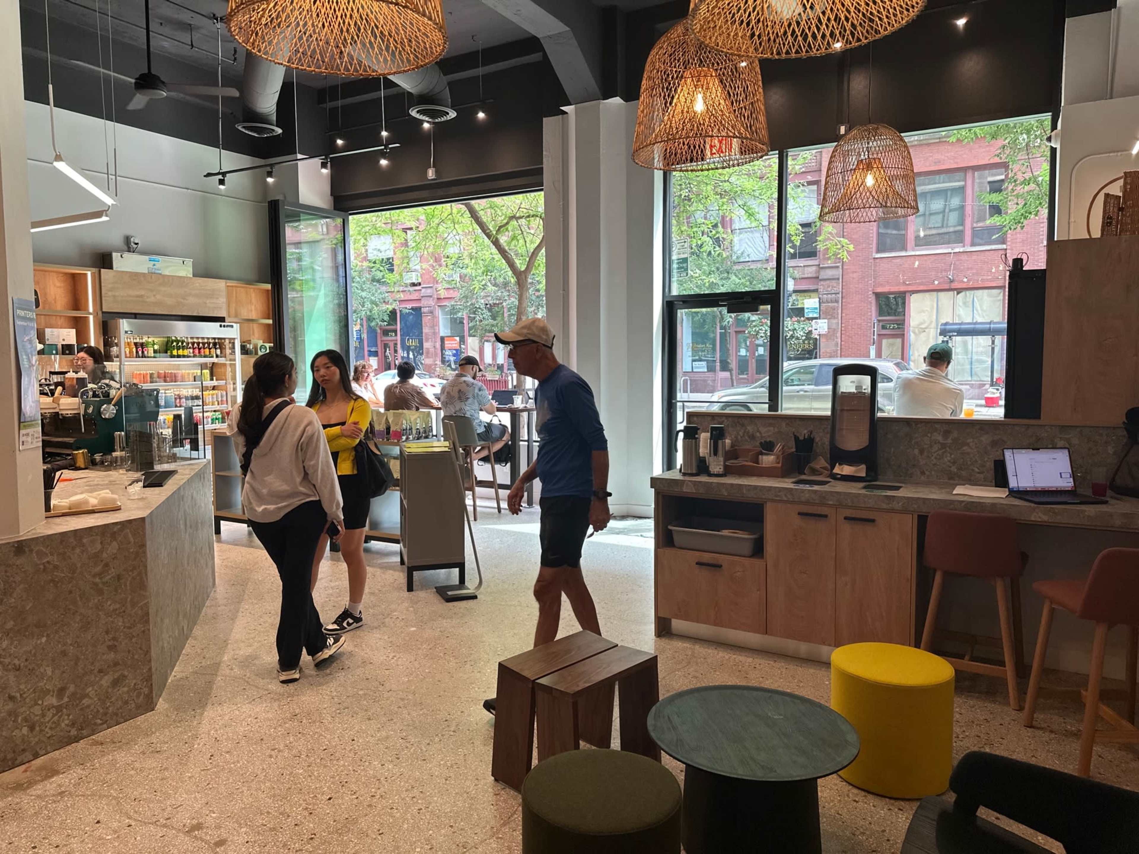 A spacious café features modern decor with large windows overlooking the street, where patrons are engaged in conversation and ordering drinks.