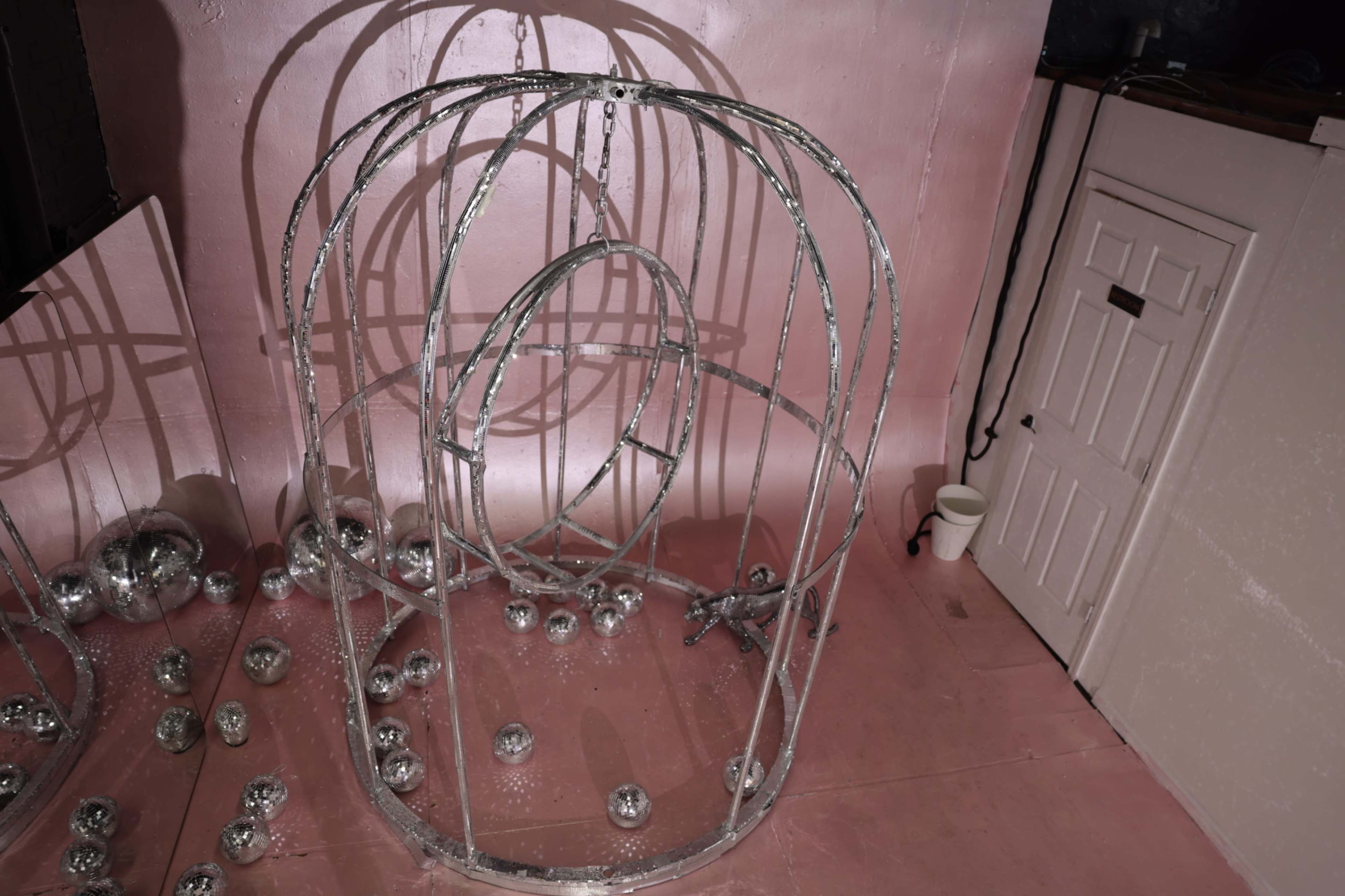 The image shows a wire-framed structure resembling a cage, surrounded by small mirrored balls, against a pink wall and floor.