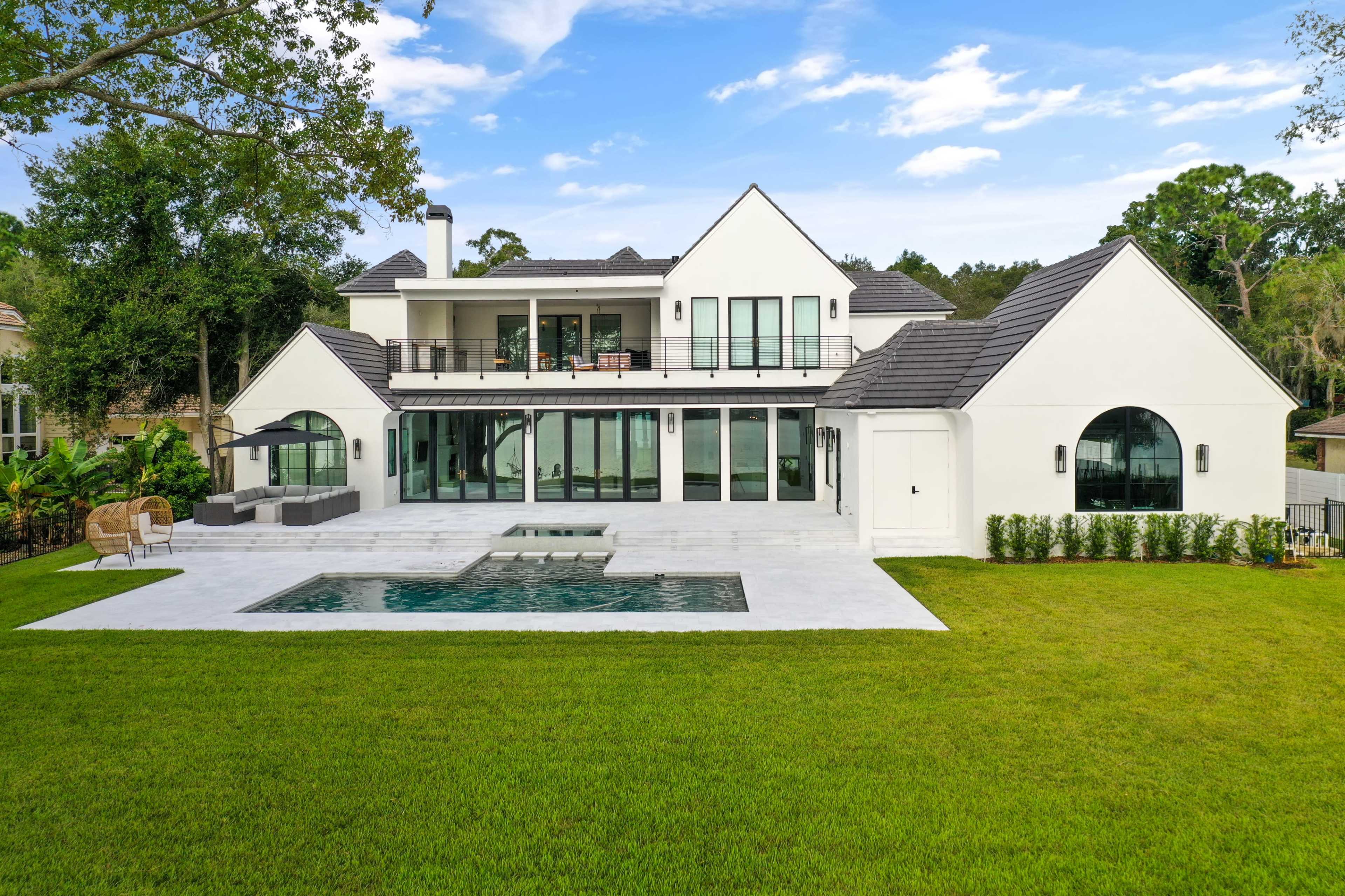 A modern two-story house with large windows and a swimming pool is set on a grassy lawn surrounded by trees.