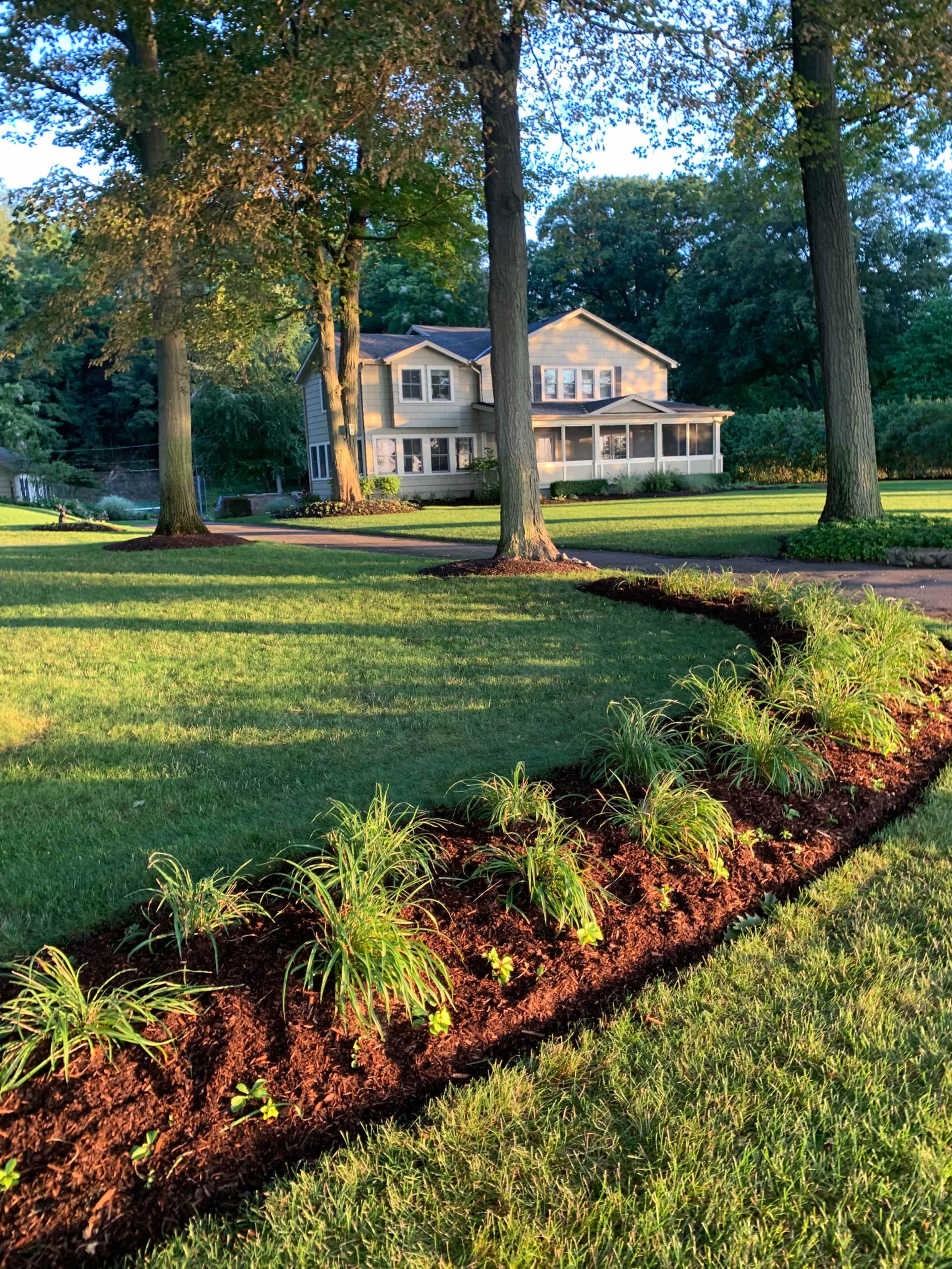 A landscaped yard features a flower bed with new plants bordered by mulch, leading up to a large house with a porch.