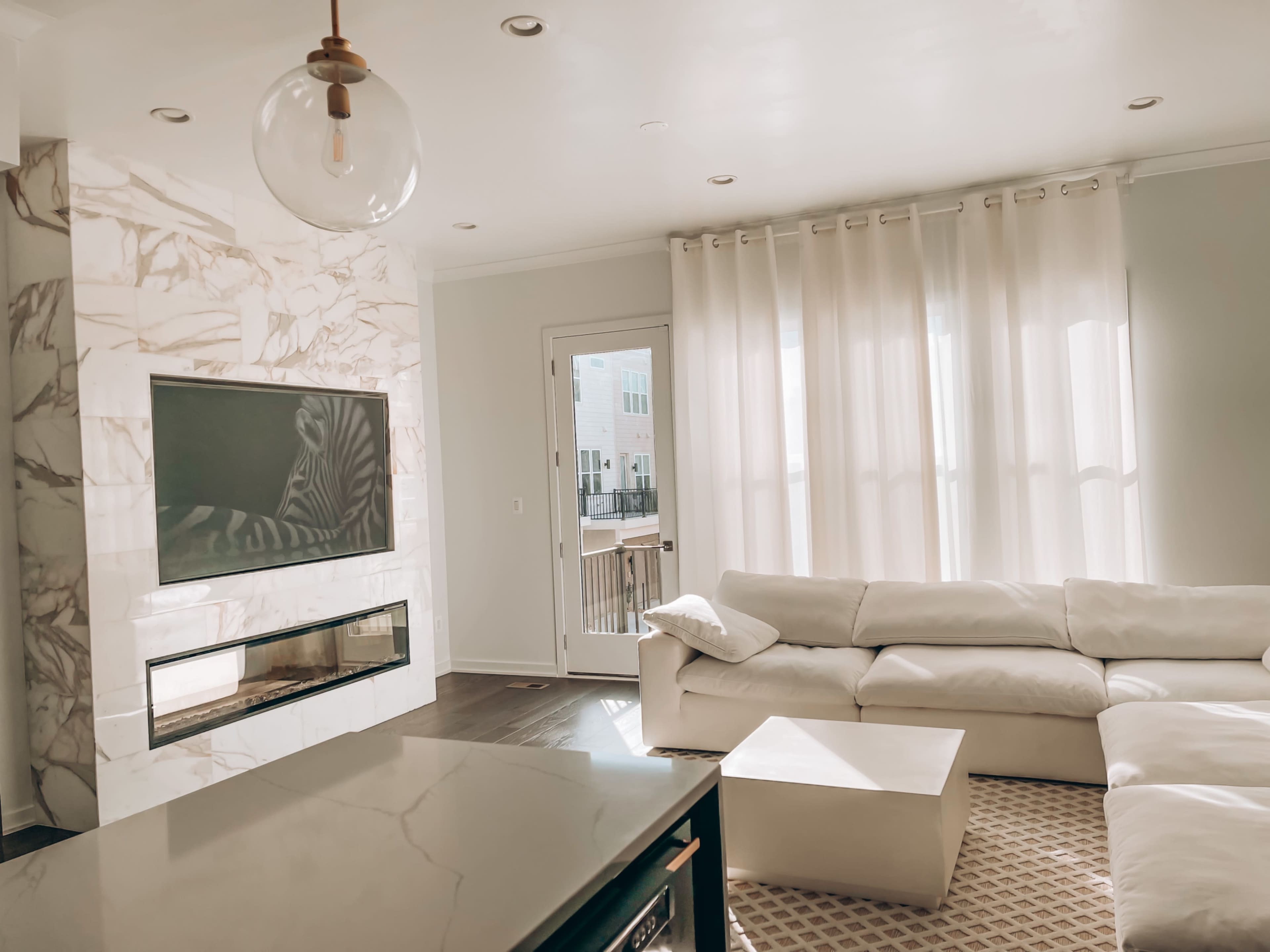 A modern living room features a marble accent wall with a flat-screen TV, large windows draped with sheer curtains, and a white sectional sofa.