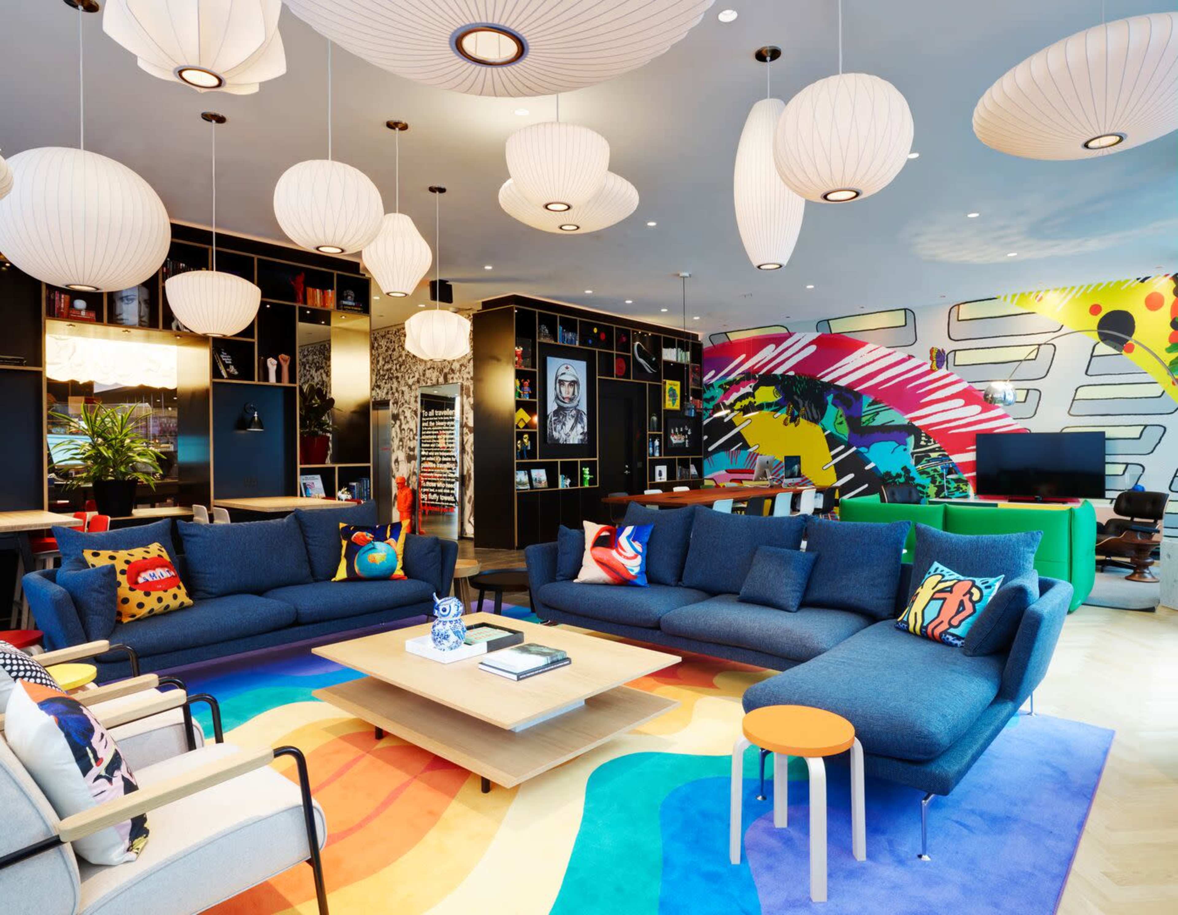 A modern living room features a large blue sectional sofa, a colorful rug, and multiple decorative pendant lights, with vibrant wall art and a bookshelf in the background.