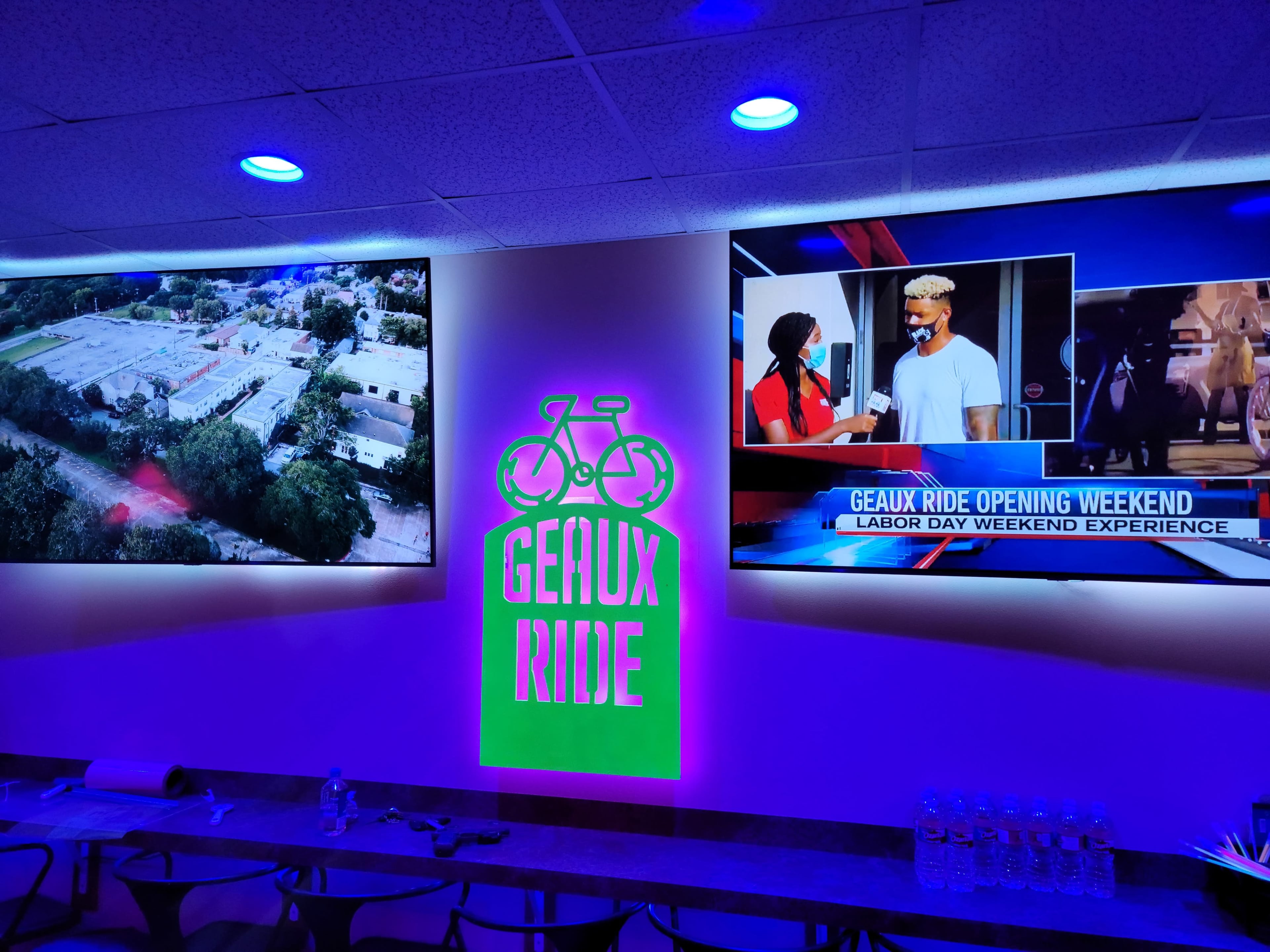 A neon sign for "Geaux Ride" next to two large screens displaying an aerial view of a location and a news segment with an interview.