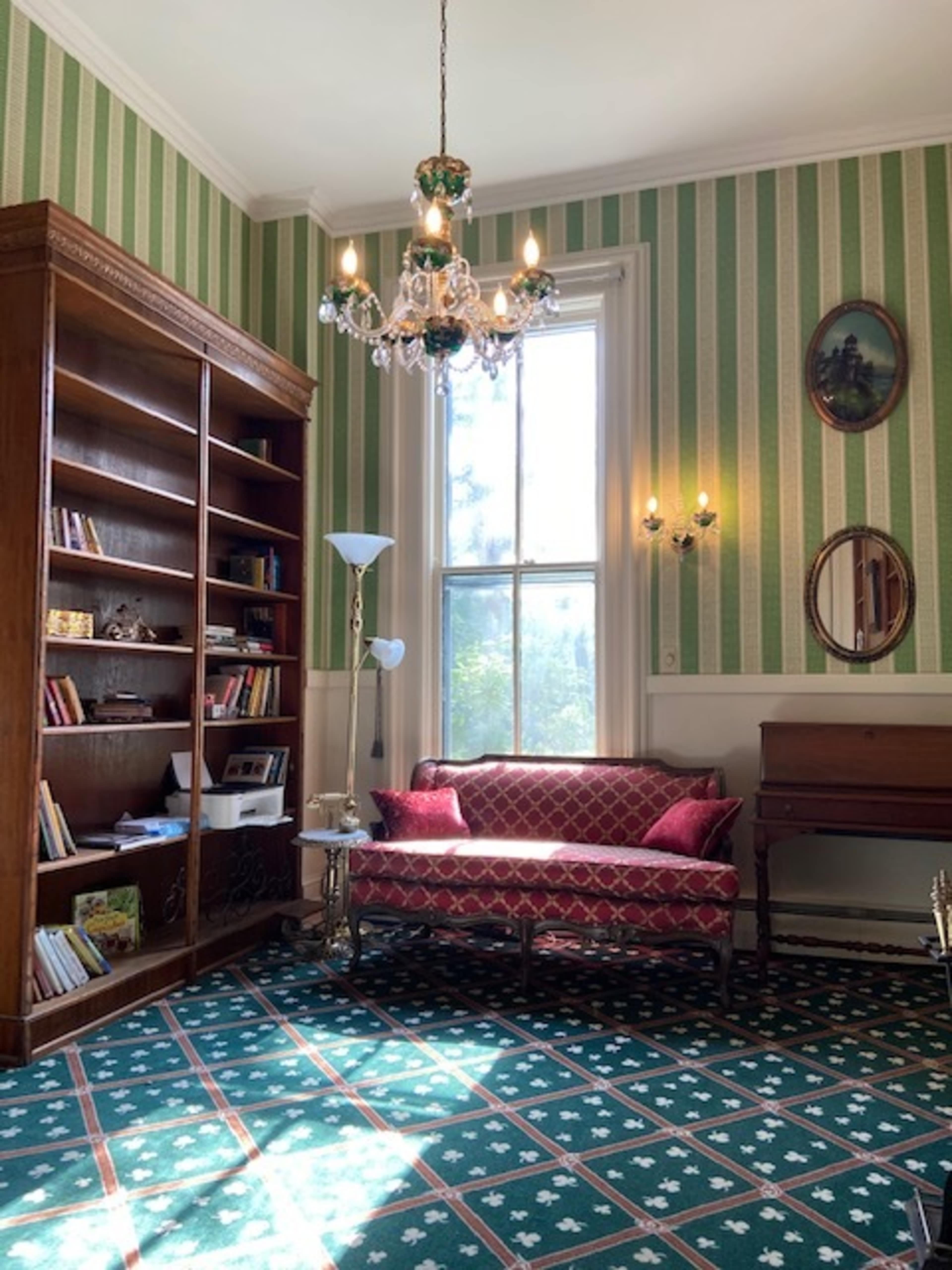 A room with striped green wallpaper, a wooden bookshelf filled with books, a red couch, a chandelier, and a window allowing natural light to enter.