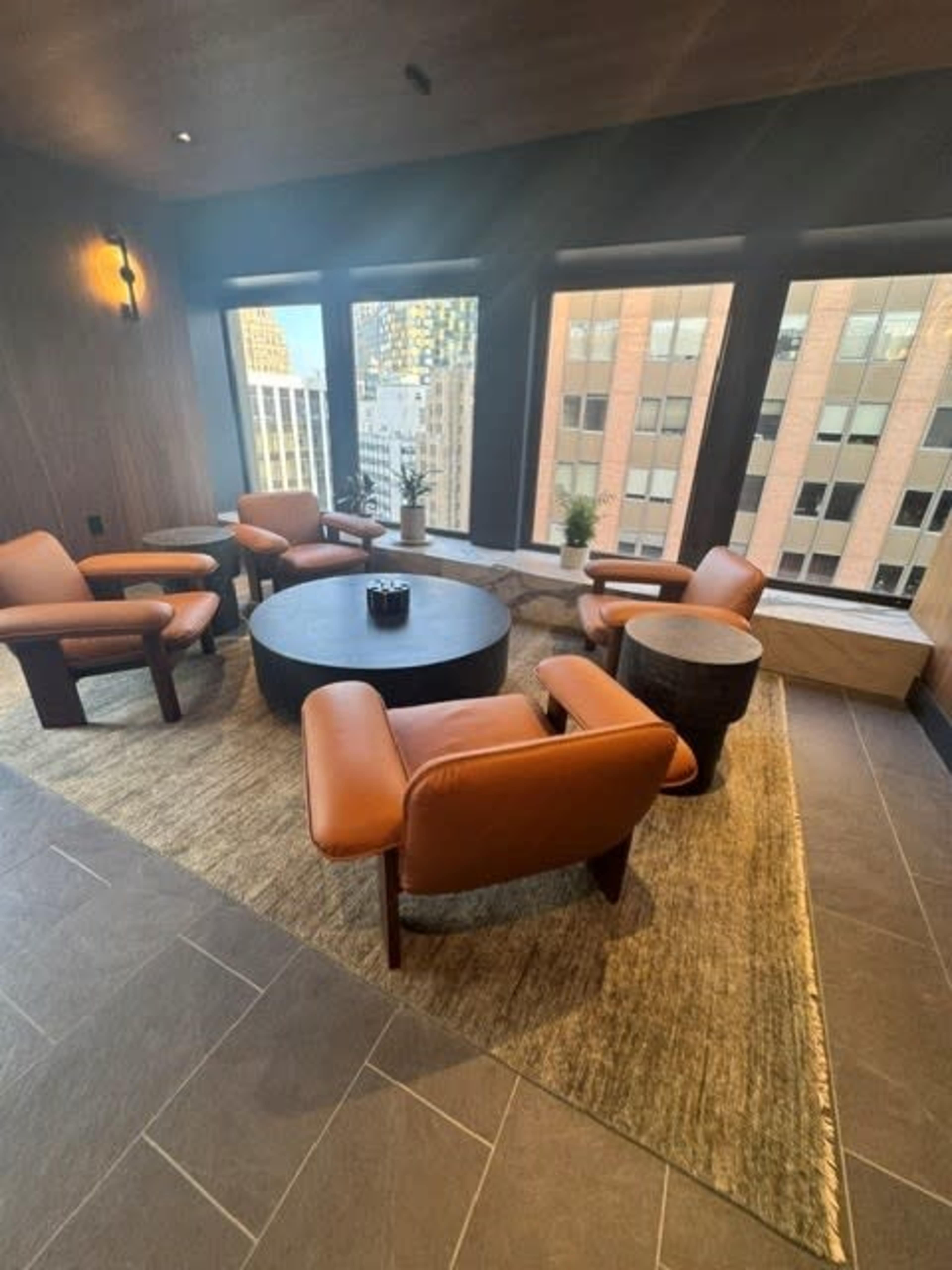 A cozy seating area features four leather chairs arranged around a round table, with large windows offering a view of a cityscape.