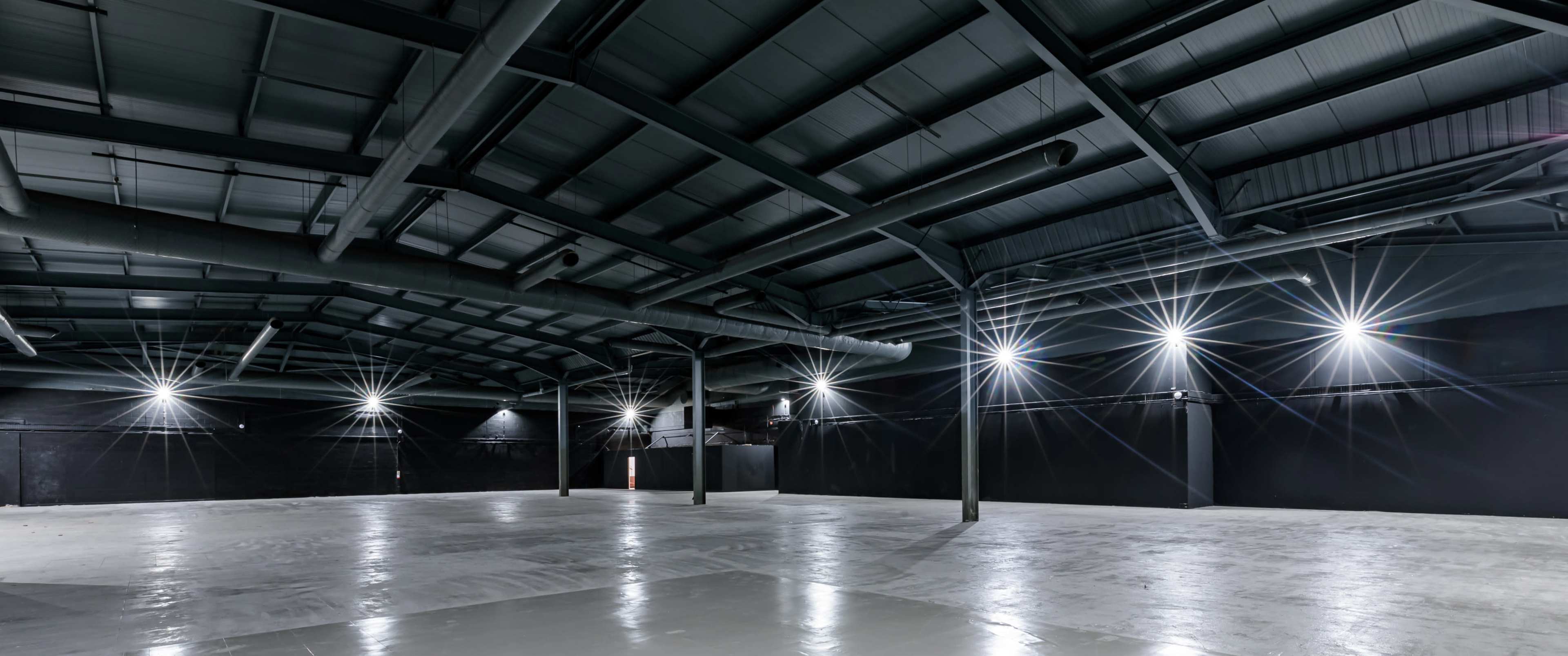 The image shows a spacious, empty industrial warehouse with a high, metal roof and bright lighting.