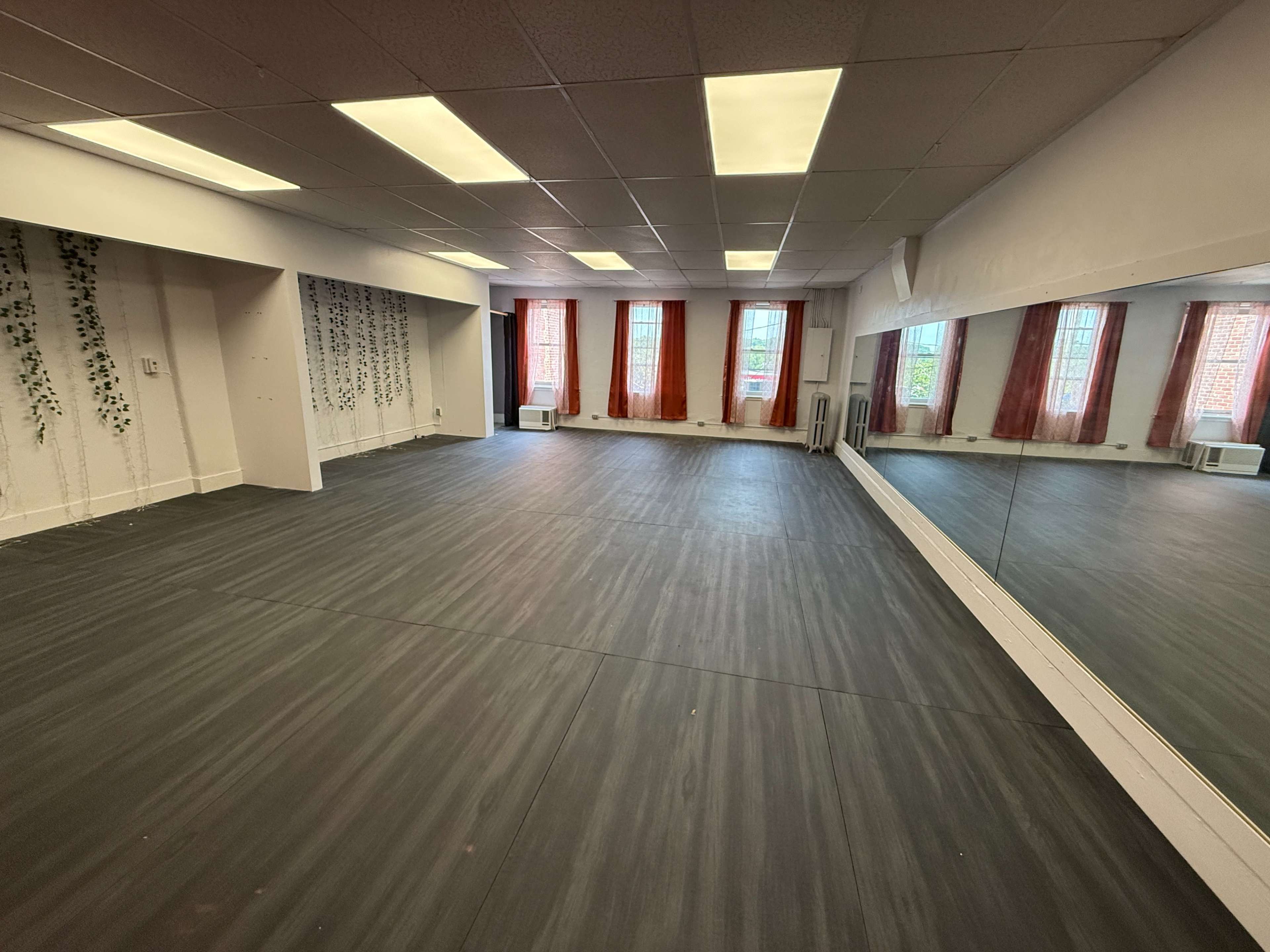 The image shows a spacious room with wooden flooring, large windows covered in sheer curtains, and a full-length mirror on one wall.