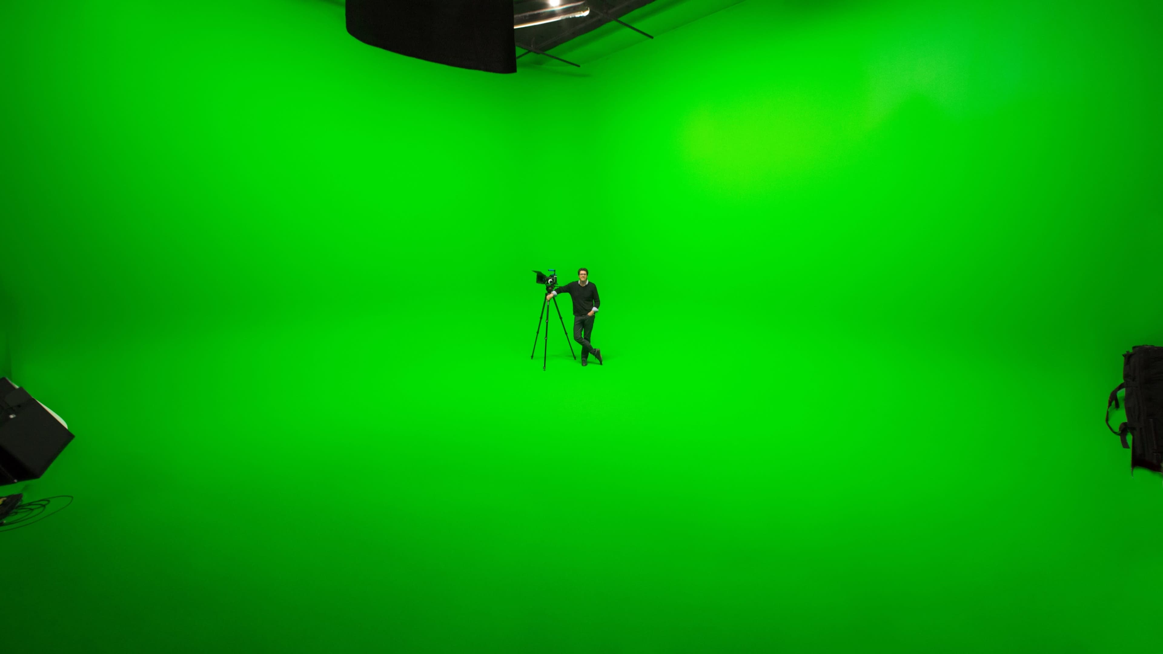 A person stands on a green backdrop in a studio, with a camera on a tripod positioned nearby.