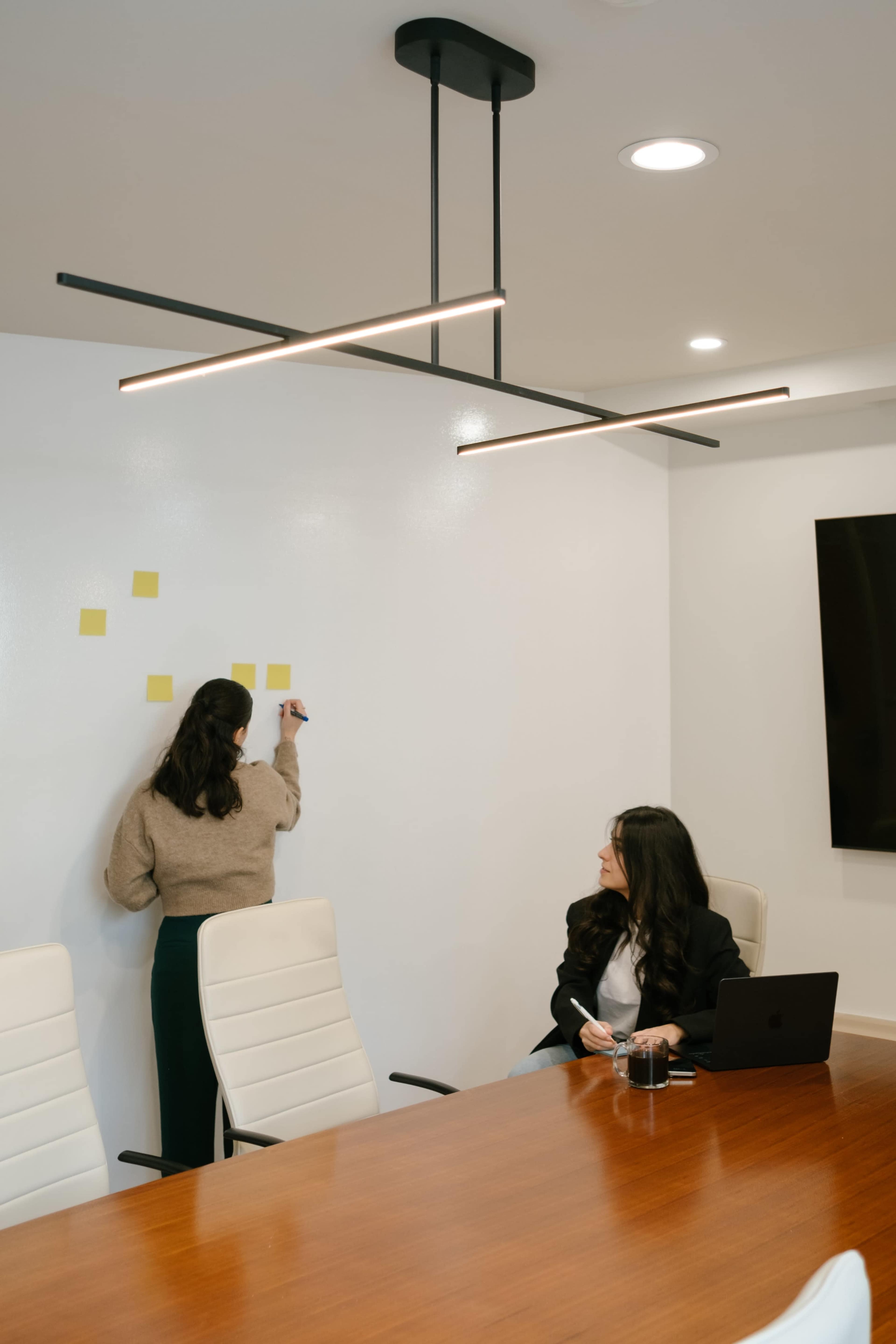 A woman writes on a whiteboard with sticky notes while another woman sits at a conference table with a laptop.