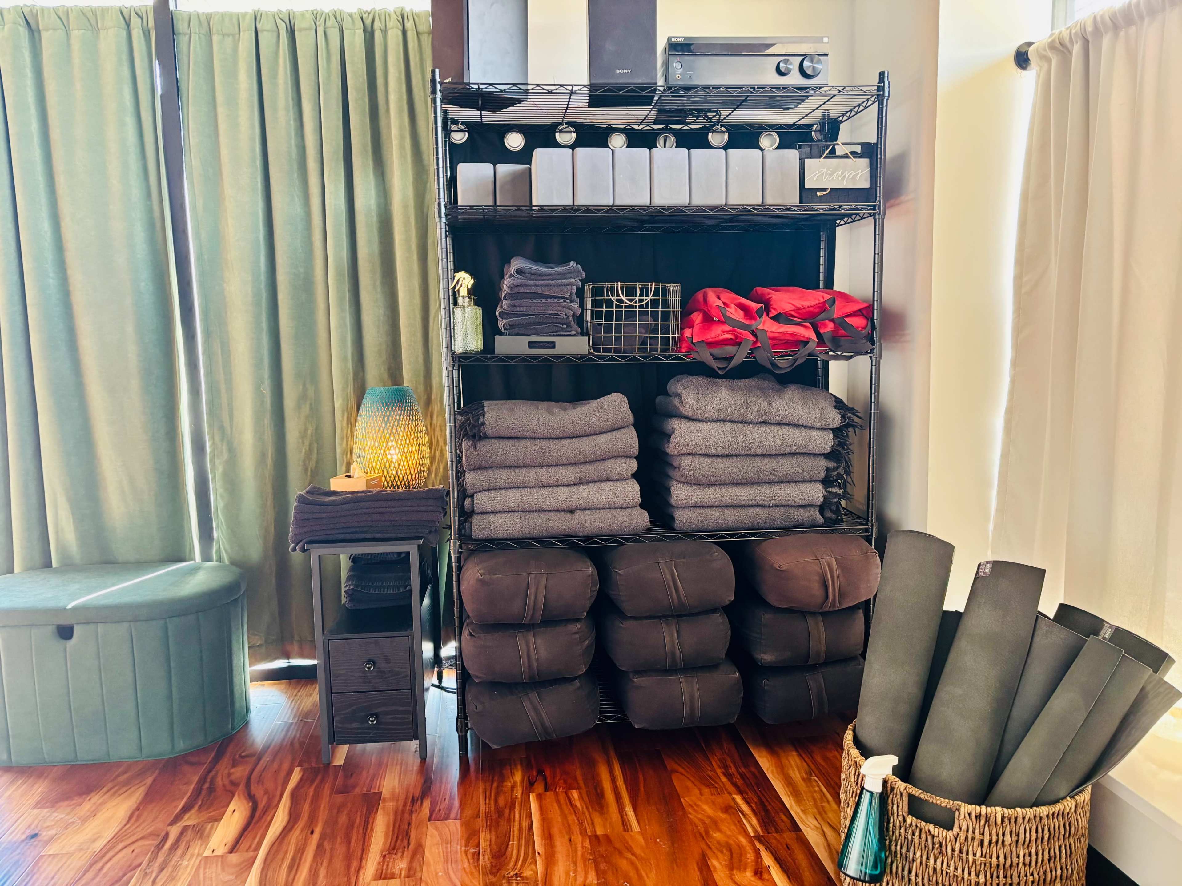 The image shows a neatly organized shelving unit filled with folded towels and blankets, accompanied by decorative items and storage containers, set against green curtains and wooden flooring.