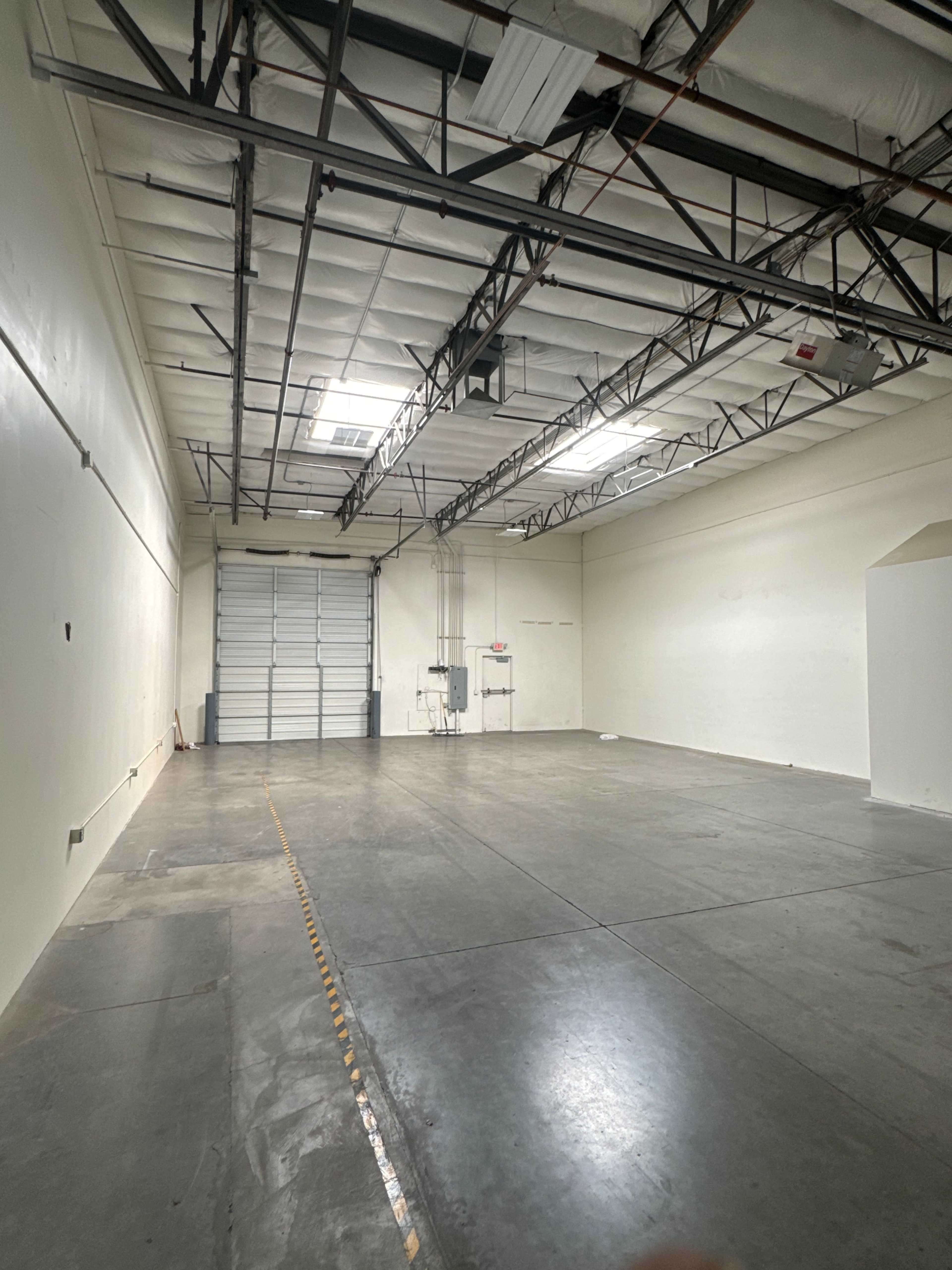 The image shows a spacious, empty industrial room with a concrete floor, high ceiling, and a large roll-up door at one end.