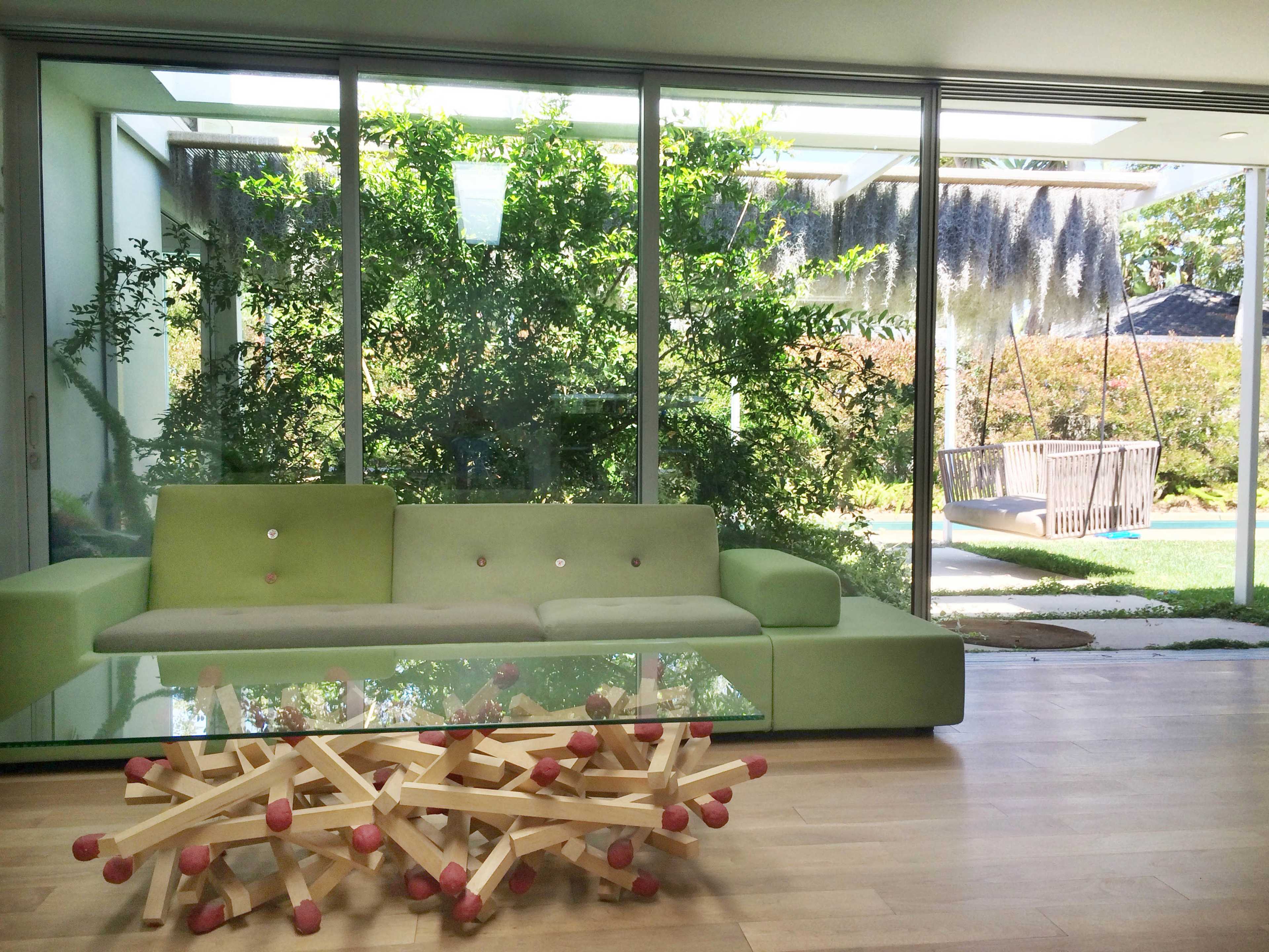 A modern living room features a green sofa and unique coffee table, with large windows overlooking a garden with trees and a swing.