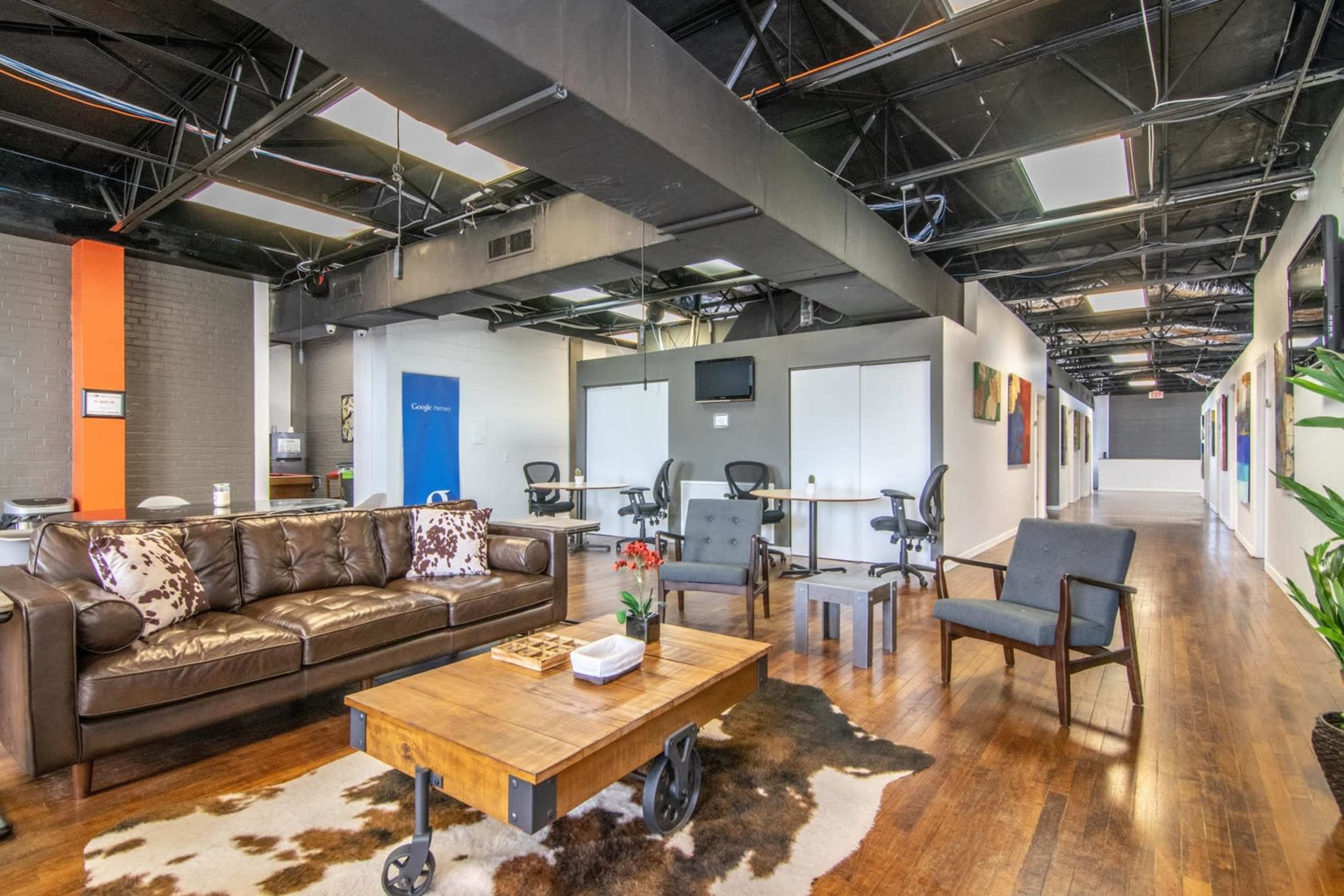 A modern office interior with a brown leather couch, wooden coffee table, and a hallway lined with colorful artwork and meeting rooms.