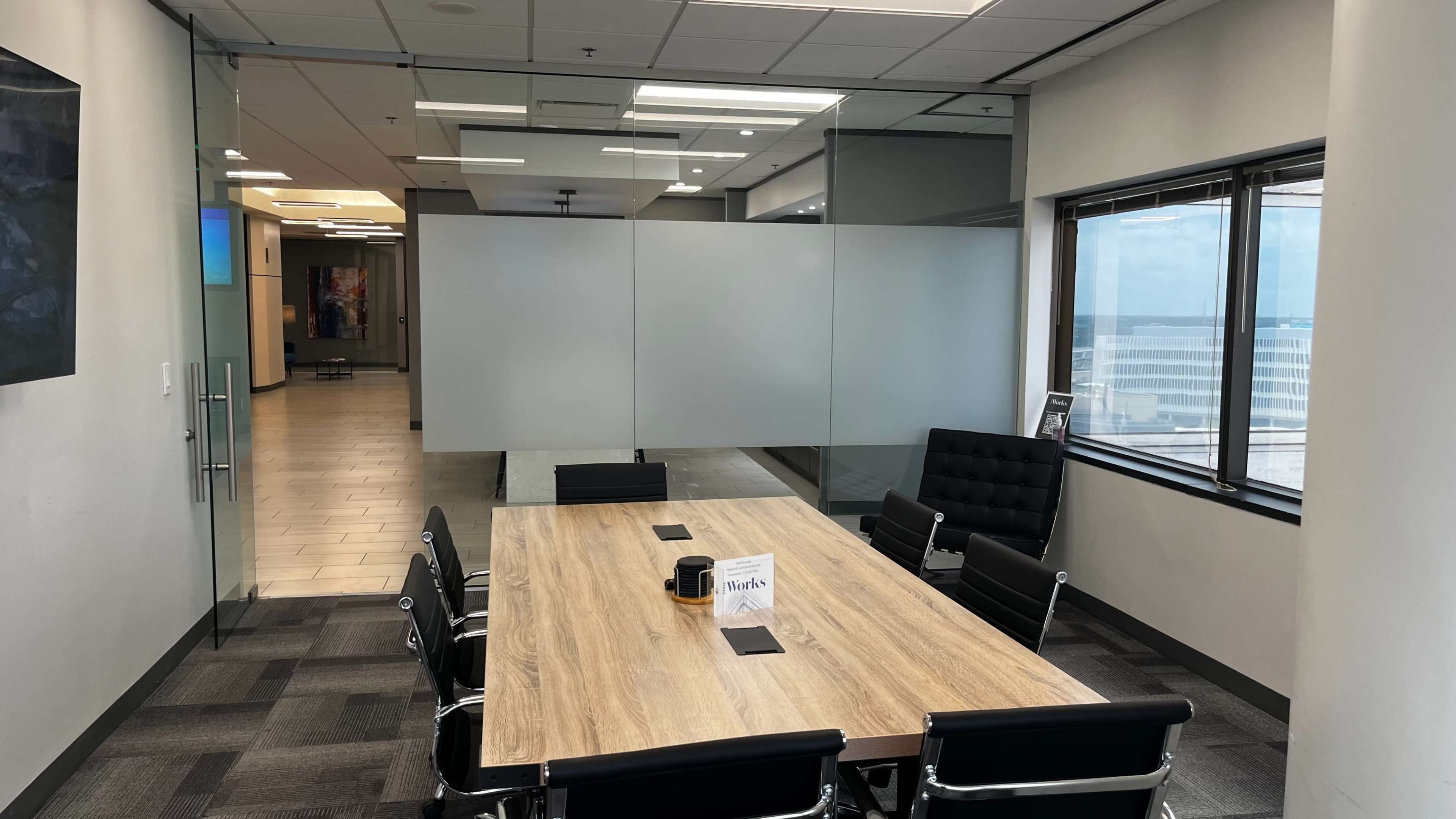 A large conference table with black chairs is situated in a modern office space, featuring frosted glass partitions and large windows overlooking a cityscape.