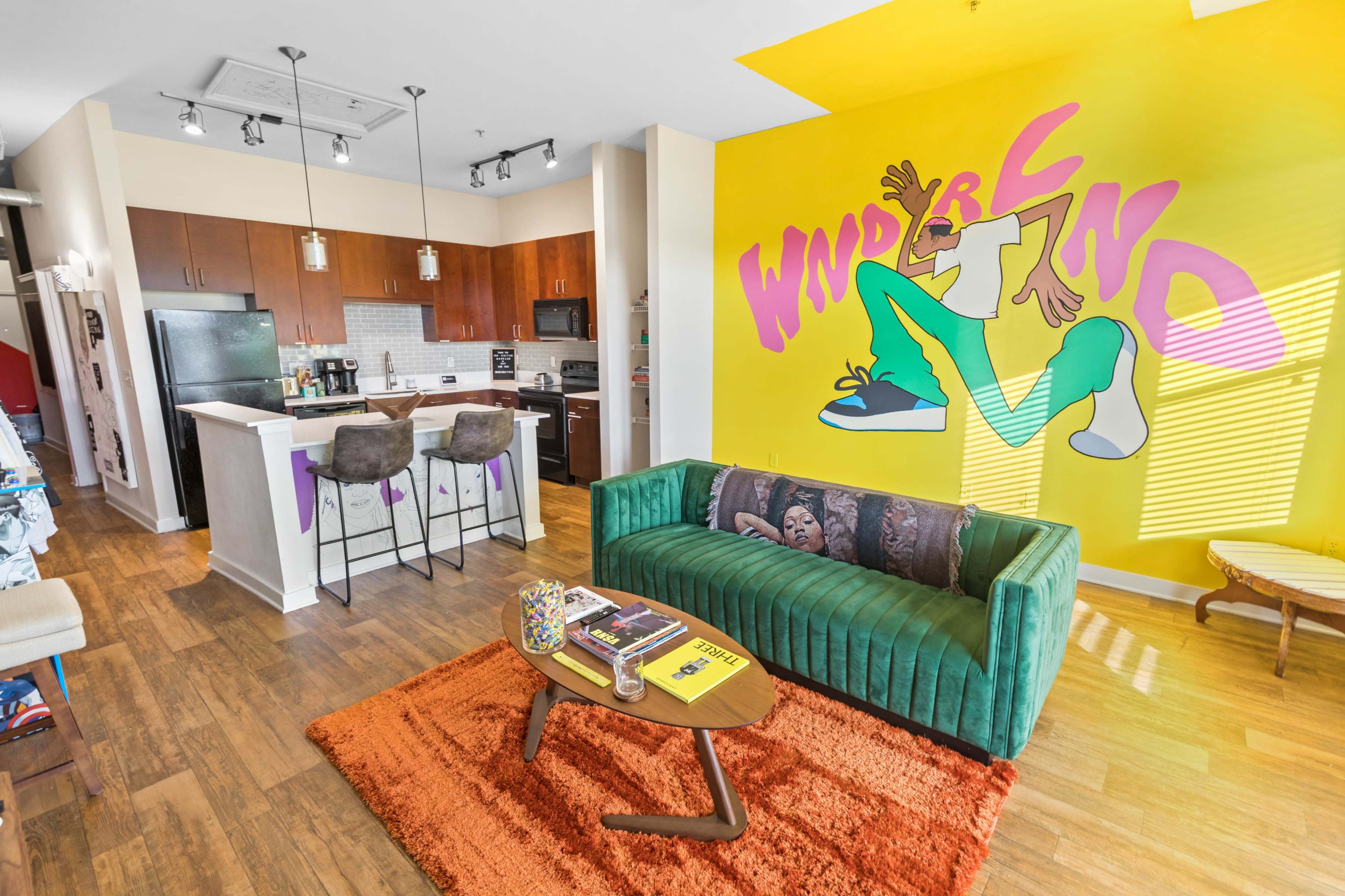 A modern apartment living area with a green couch, a wooden coffee table, and a kitchen space featuring dark cabinets and stainless steel appliances, highlighted by a bright yellow wall with cartoon-style lettering.