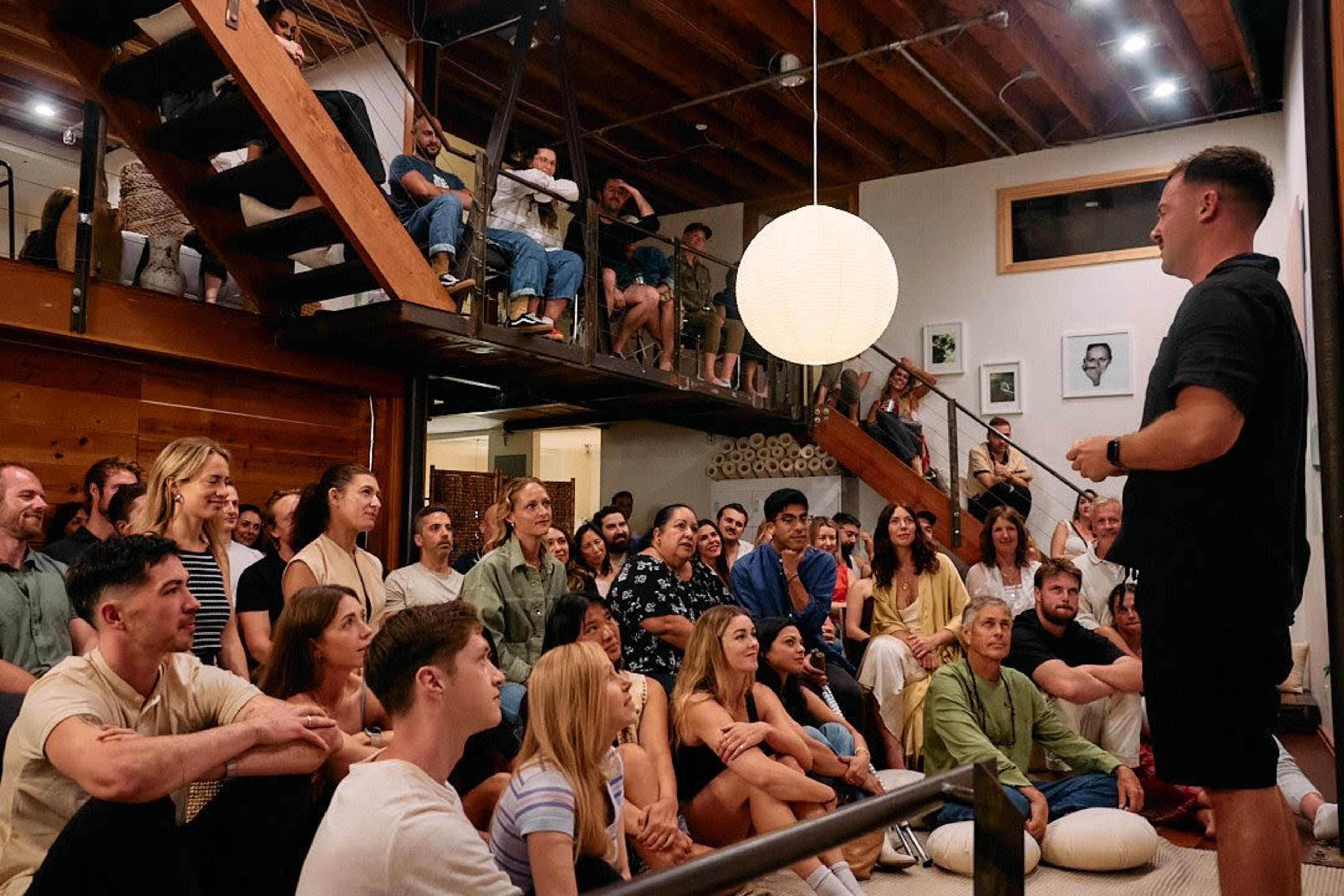 A speaker addresses an audience seated on the floor inside a well-lit, wooden interior space with stairs and artwork in the background.