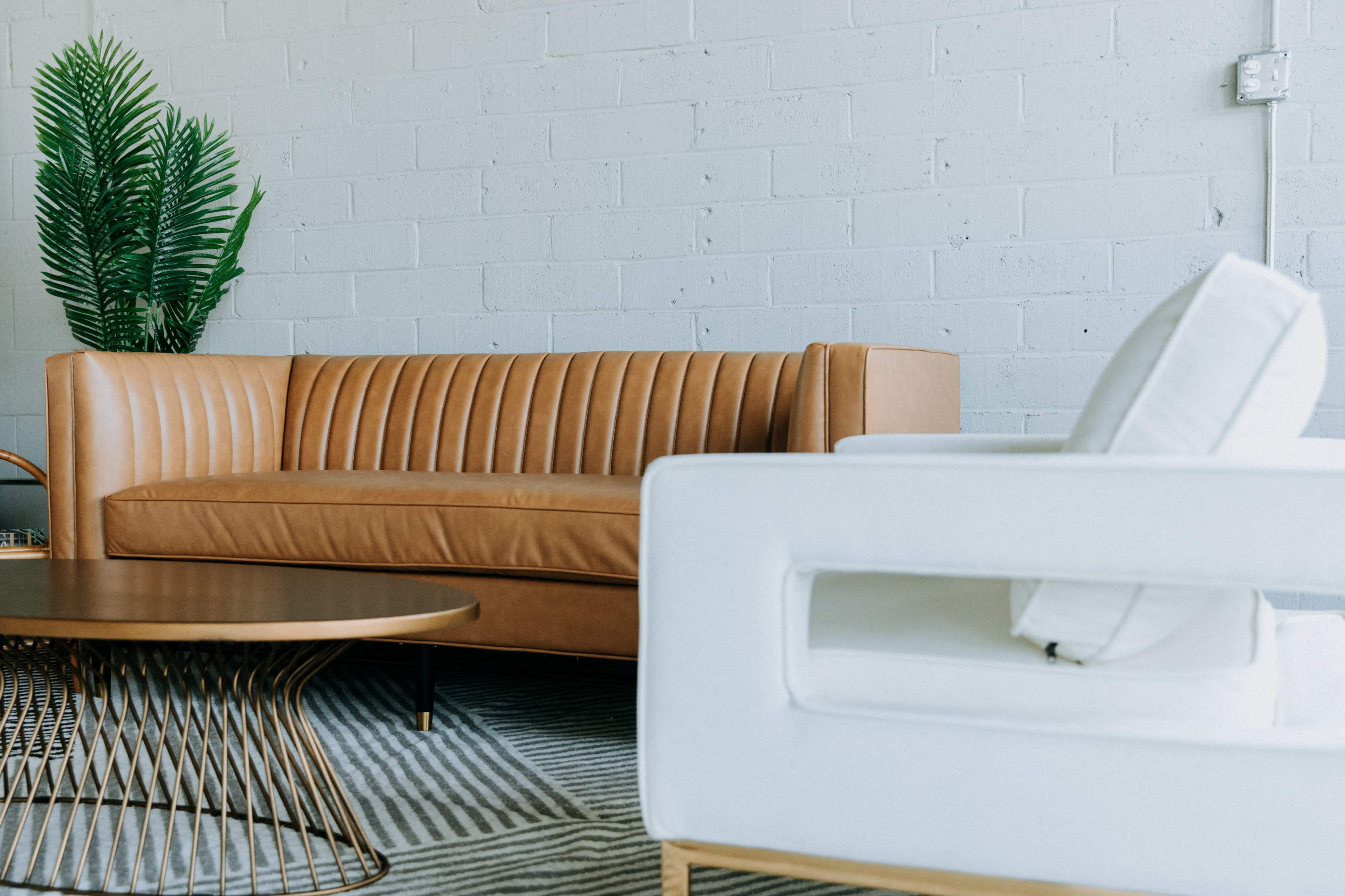 A tan leather sofa and a white armchair are positioned in a modern living area featuring a round coffee table and a potted plant against a light gray wall.