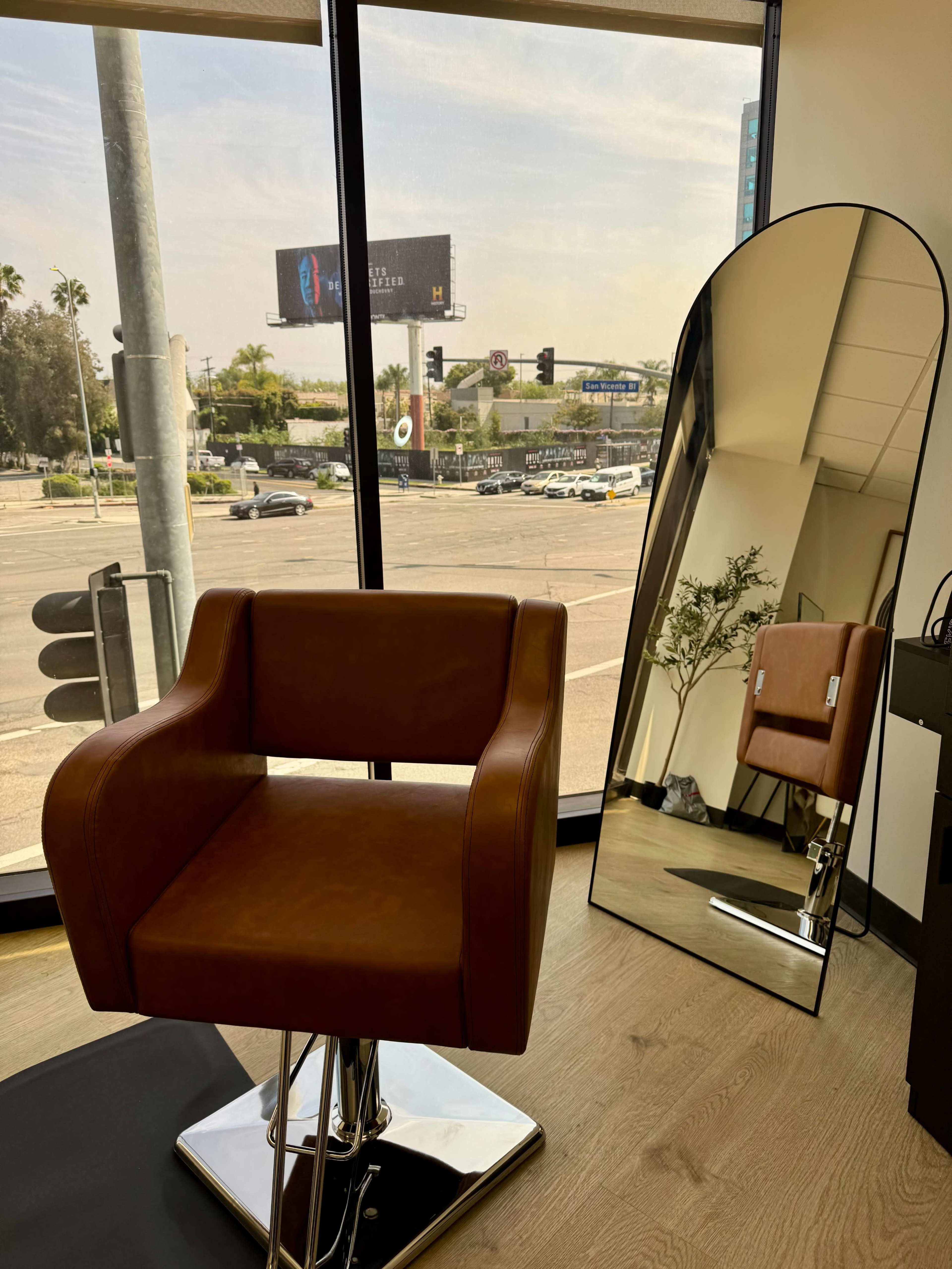 A brown salon chair is positioned in front of a large mirror, with a view of a busy street and a billboard visible through the window.
