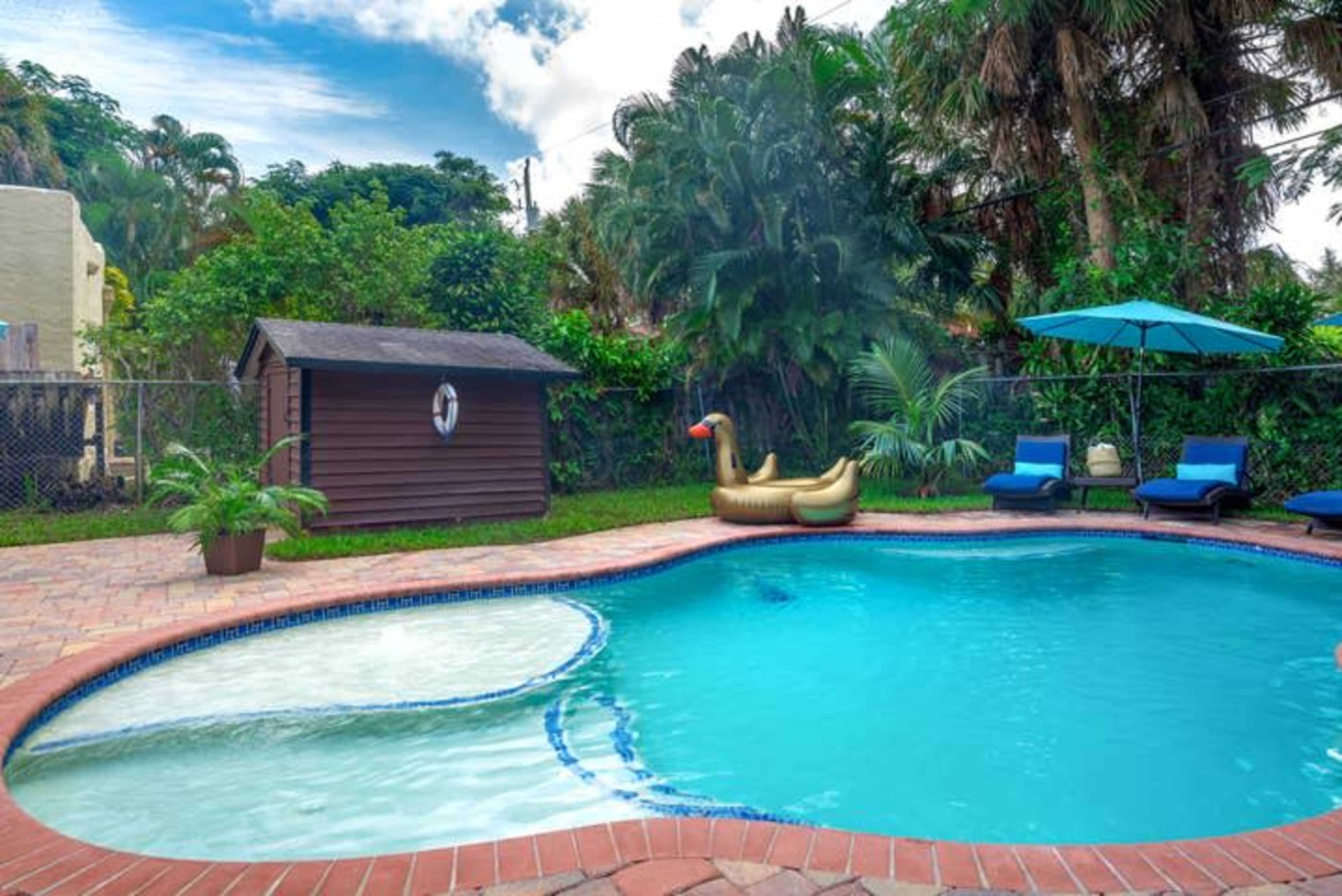 This scene features a swimming pool surrounded by lush greenery, with a large inflatable swan float and two blue lounge chairs nearby.