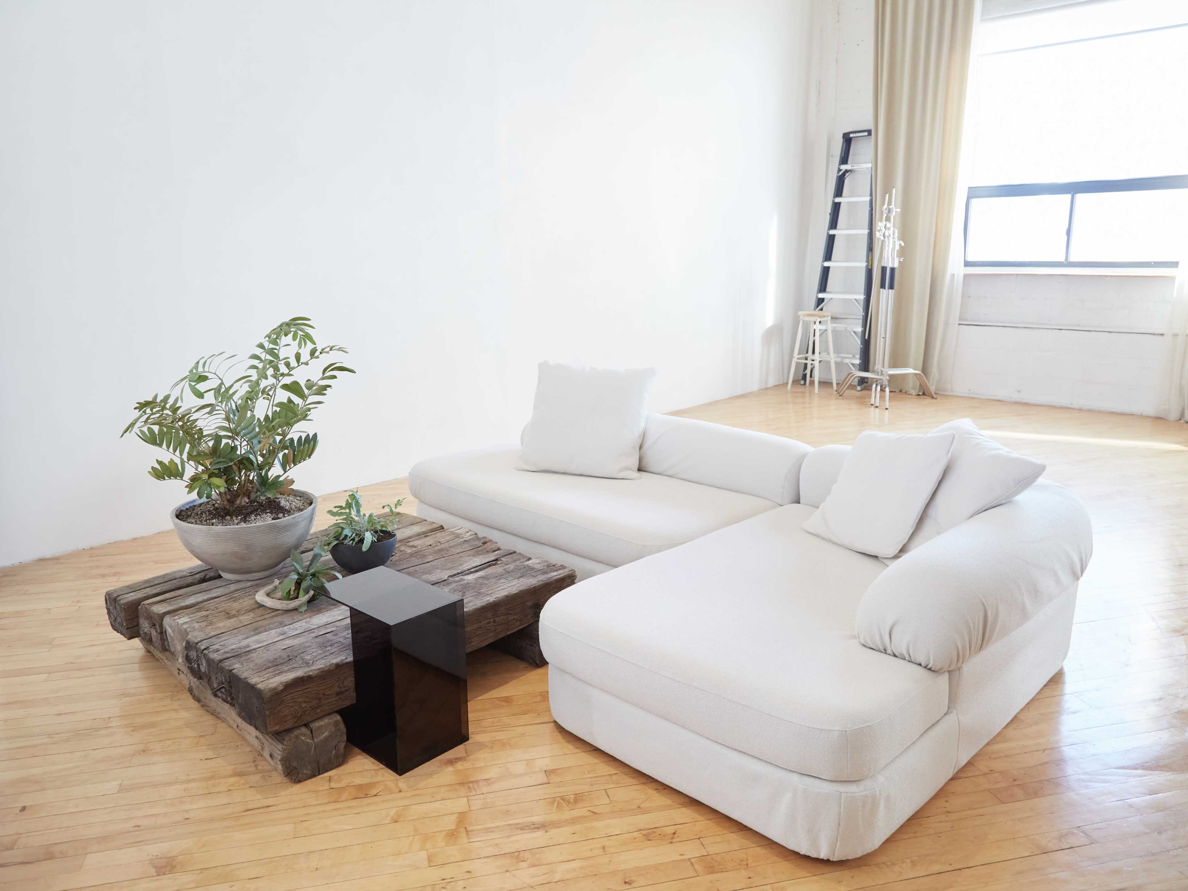 A light-colored sectional sofa and a rustic wooden coffee table are arranged in a spacious room with large windows and minimal furnishings.