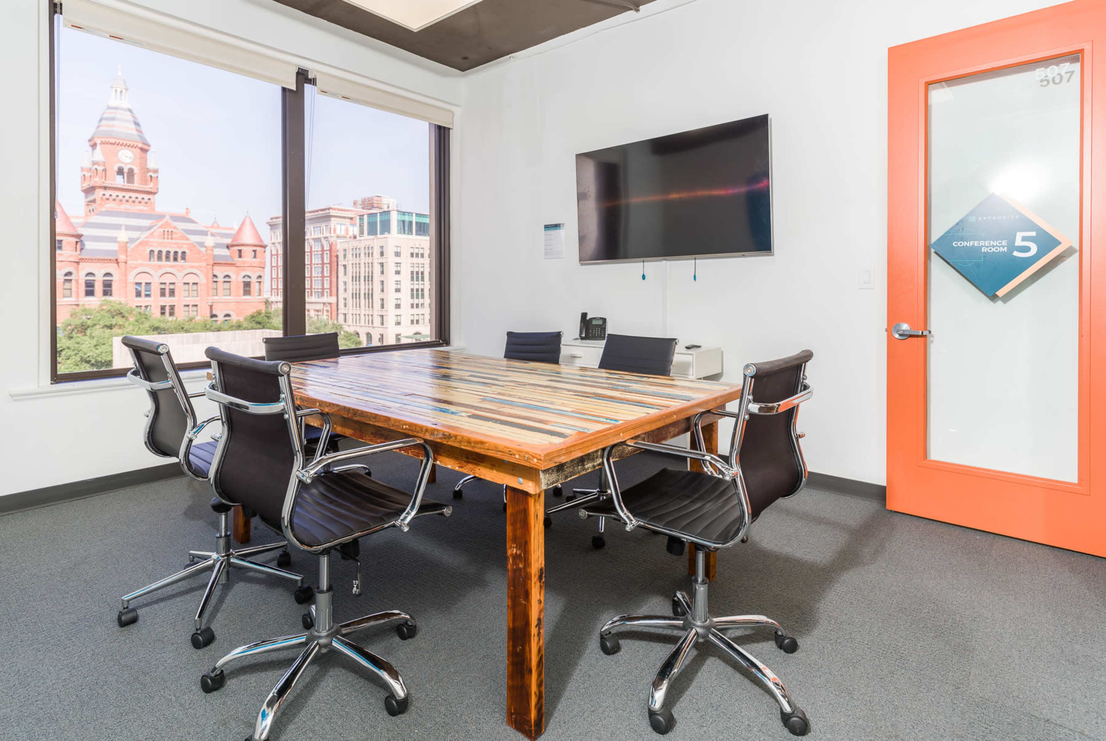 The image shows a modern conference room with a large wooden table, six black chairs, a wall-mounted TV, and a view of an exterior building through large windows.