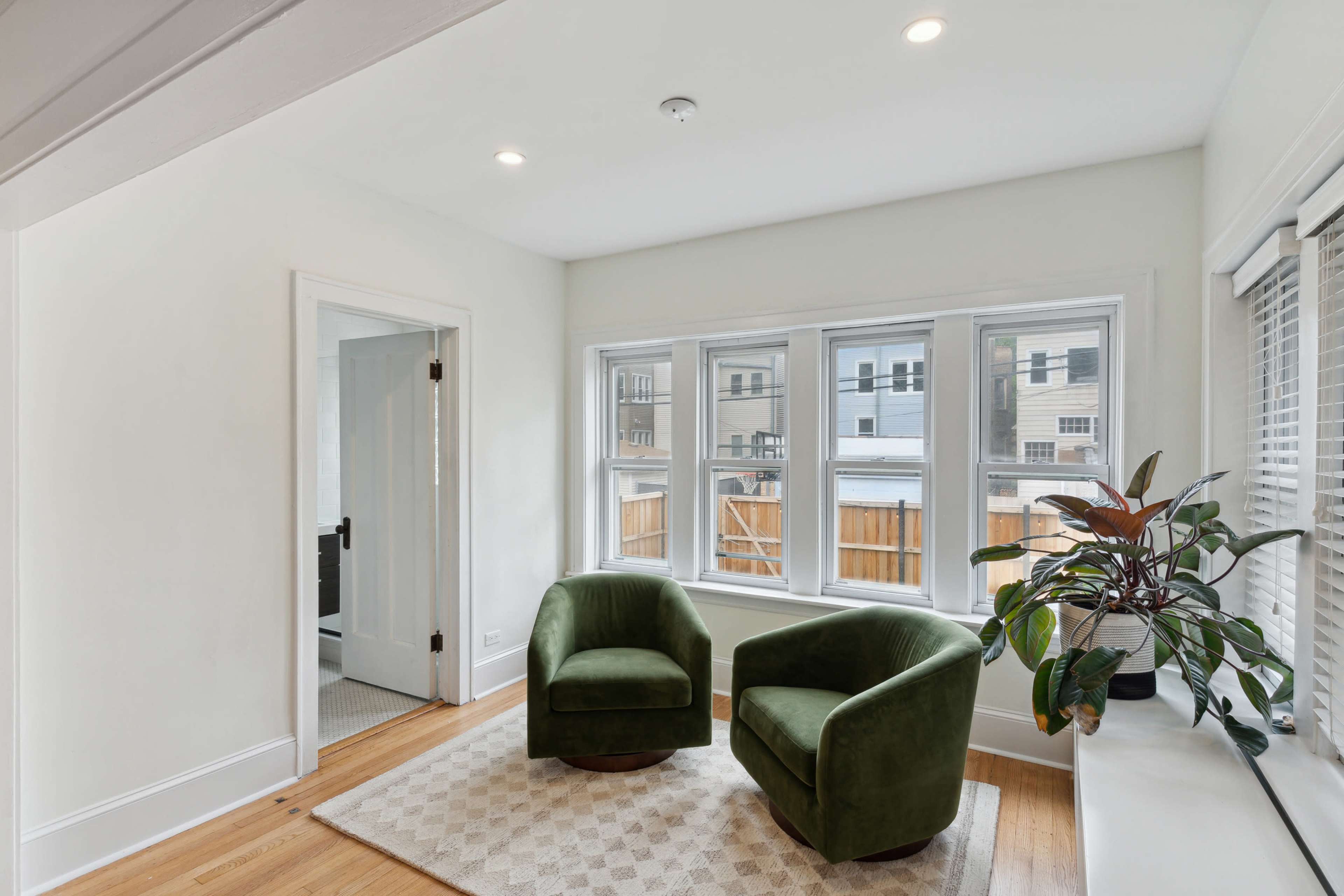 A bright corner of a room features two green armchairs, a plant, and large windows with a view of an exterior fence.