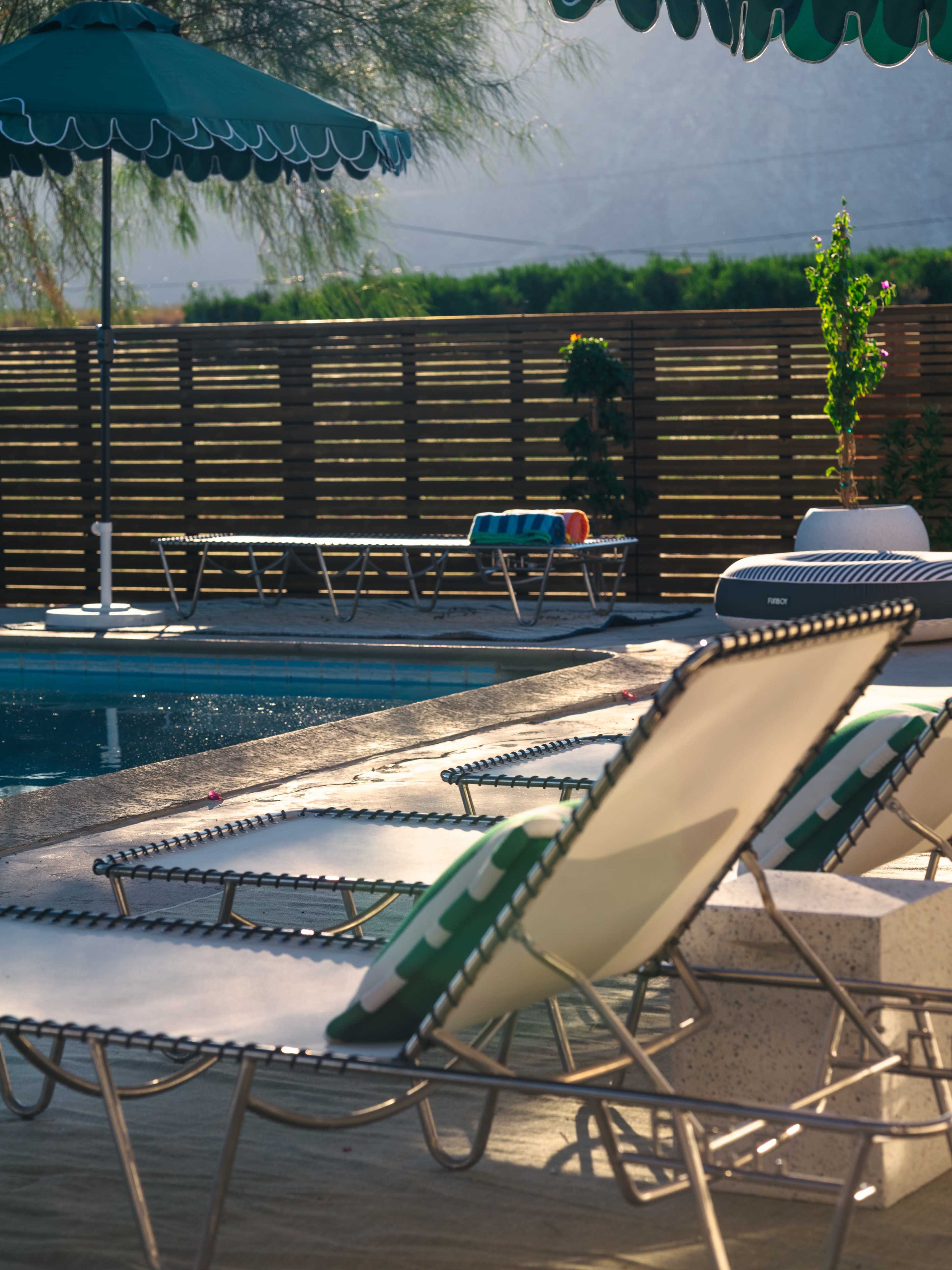 The image shows a swimming pool area with several lounge chairs, a green umbrella, and a wooden fence in the background.