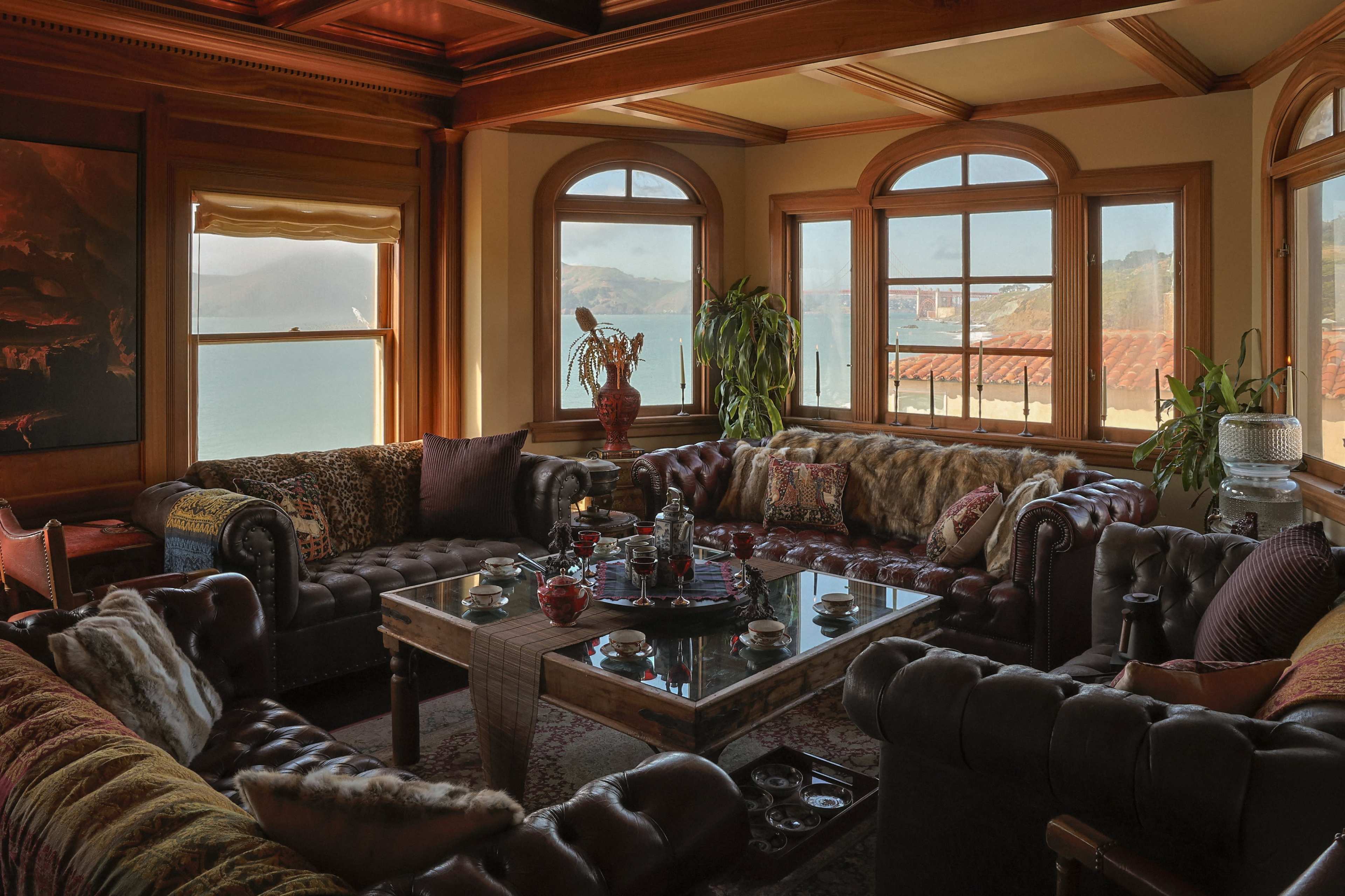 The image shows a cozy living room with leather sofas, a wooden coffee table, and large windows overlooking a body of water.