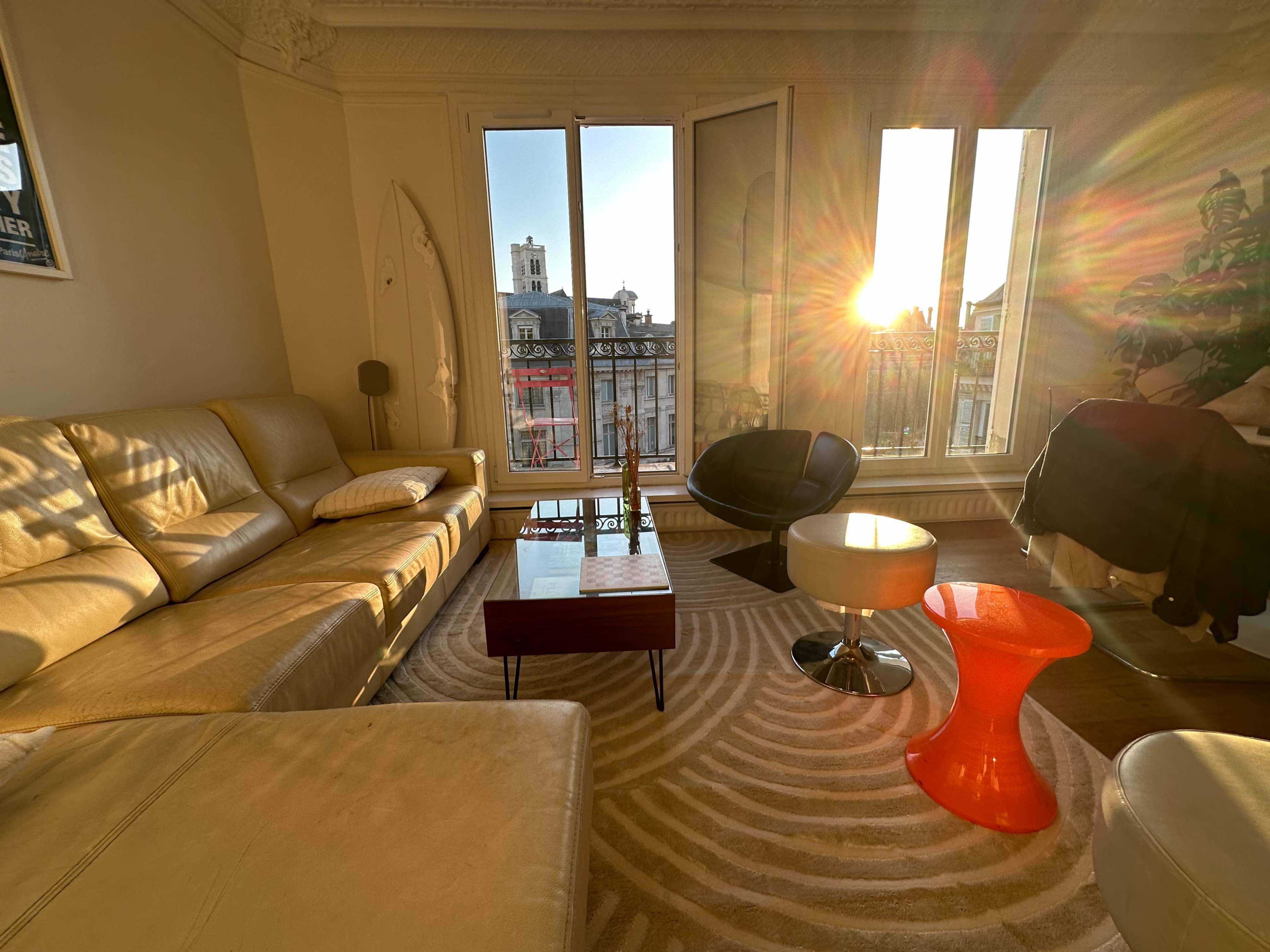 A sunlit living room features a beige leather sofa, a glass coffee table, and a vibrant red side table, with large windows opening to a cityscape.