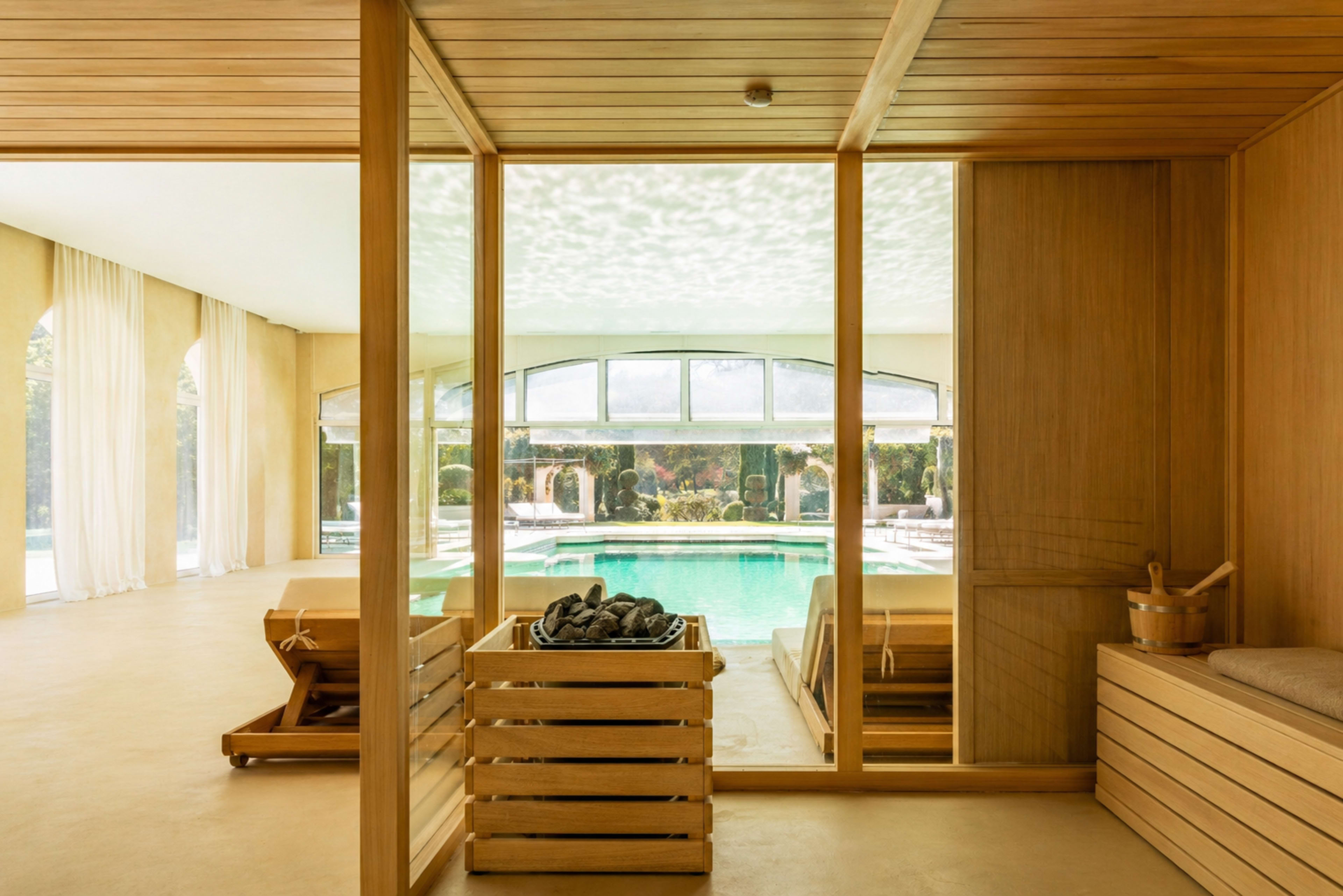 The image shows a wooden-paneled indoor space overlooking a pool with large windows and lounge chairs.