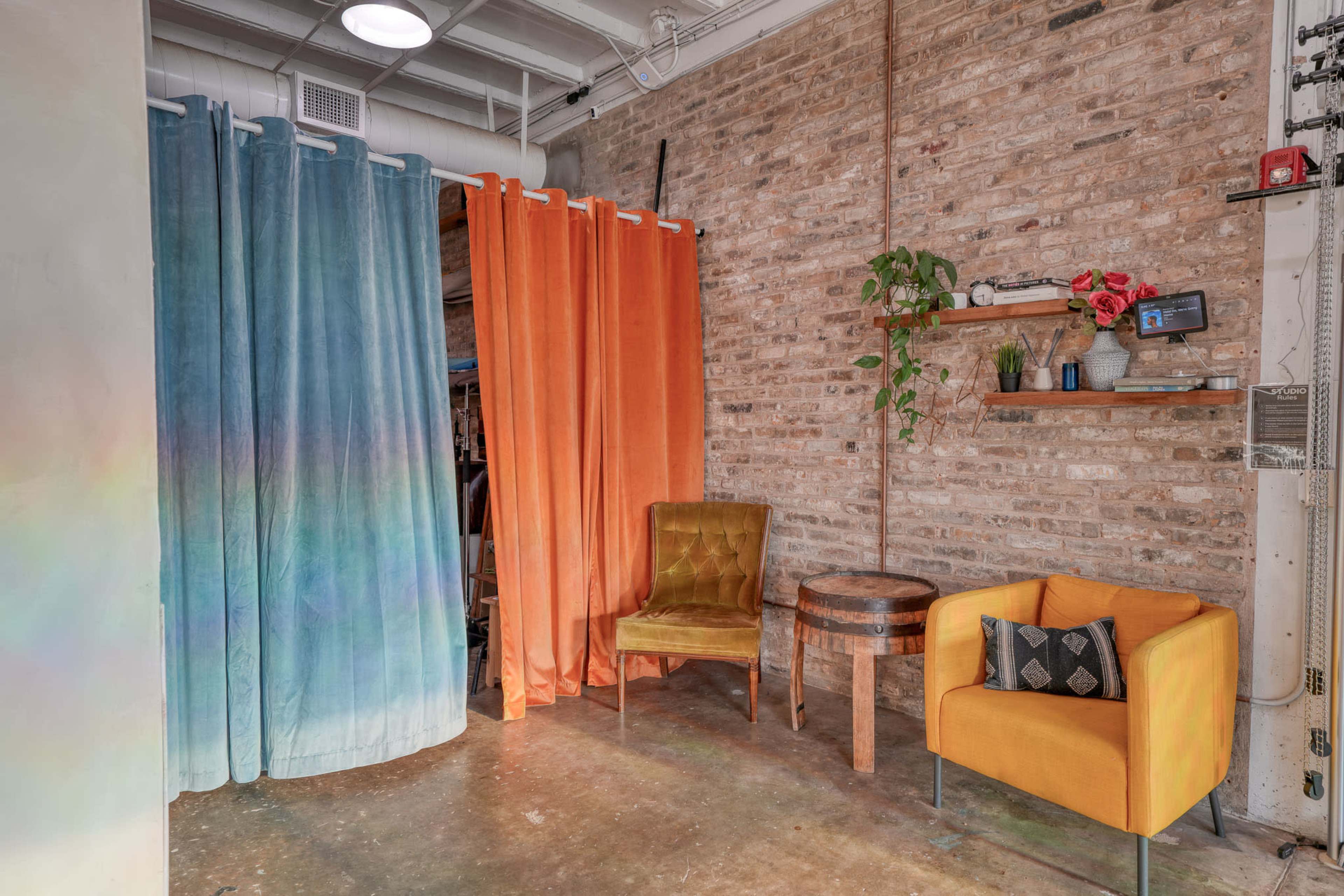 The image shows a small interior space with two colored fabric curtains, an orange chair, a yellow velvet chair, a wooden table, and a shelf with plants and flowers against a brick wall.