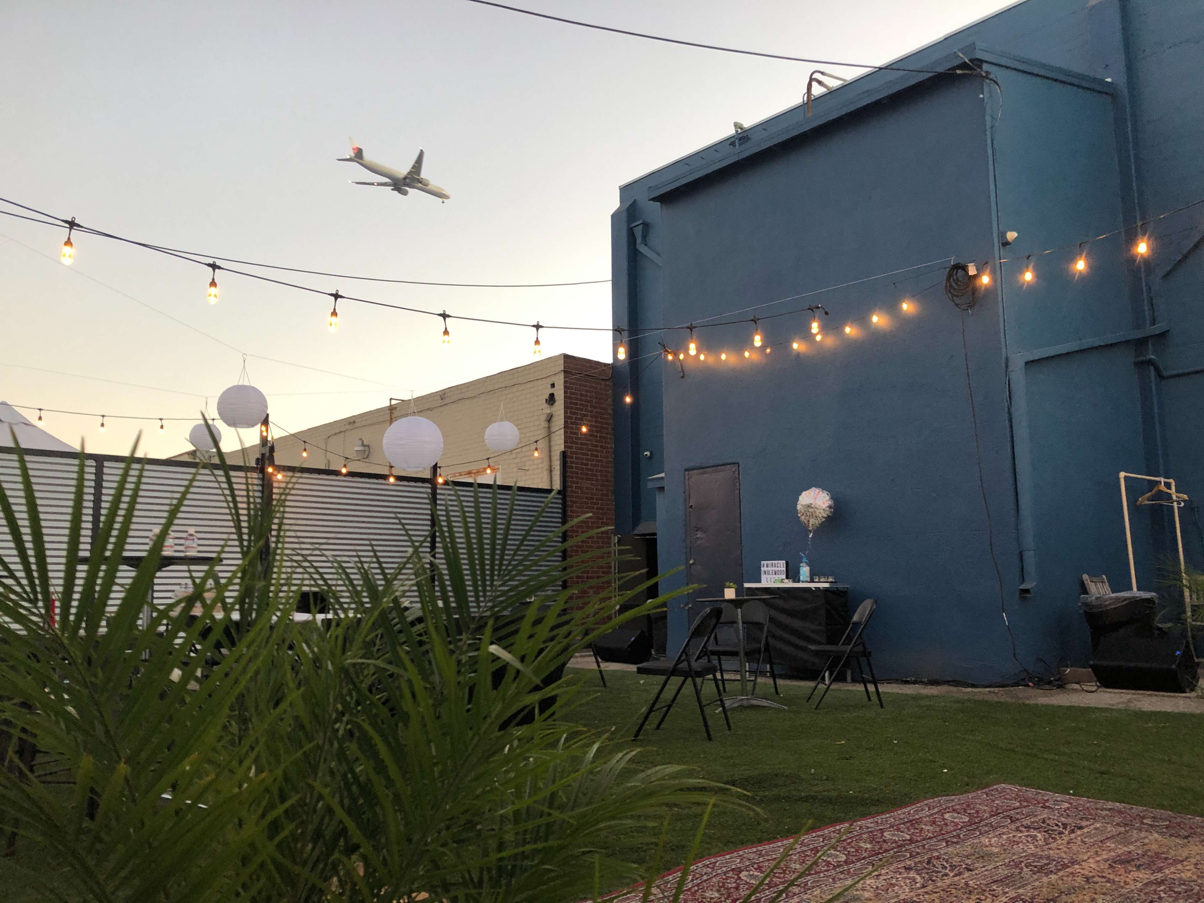 A decorated outdoor space with string lights, a table, and greenery, with an airplane flying overhead.