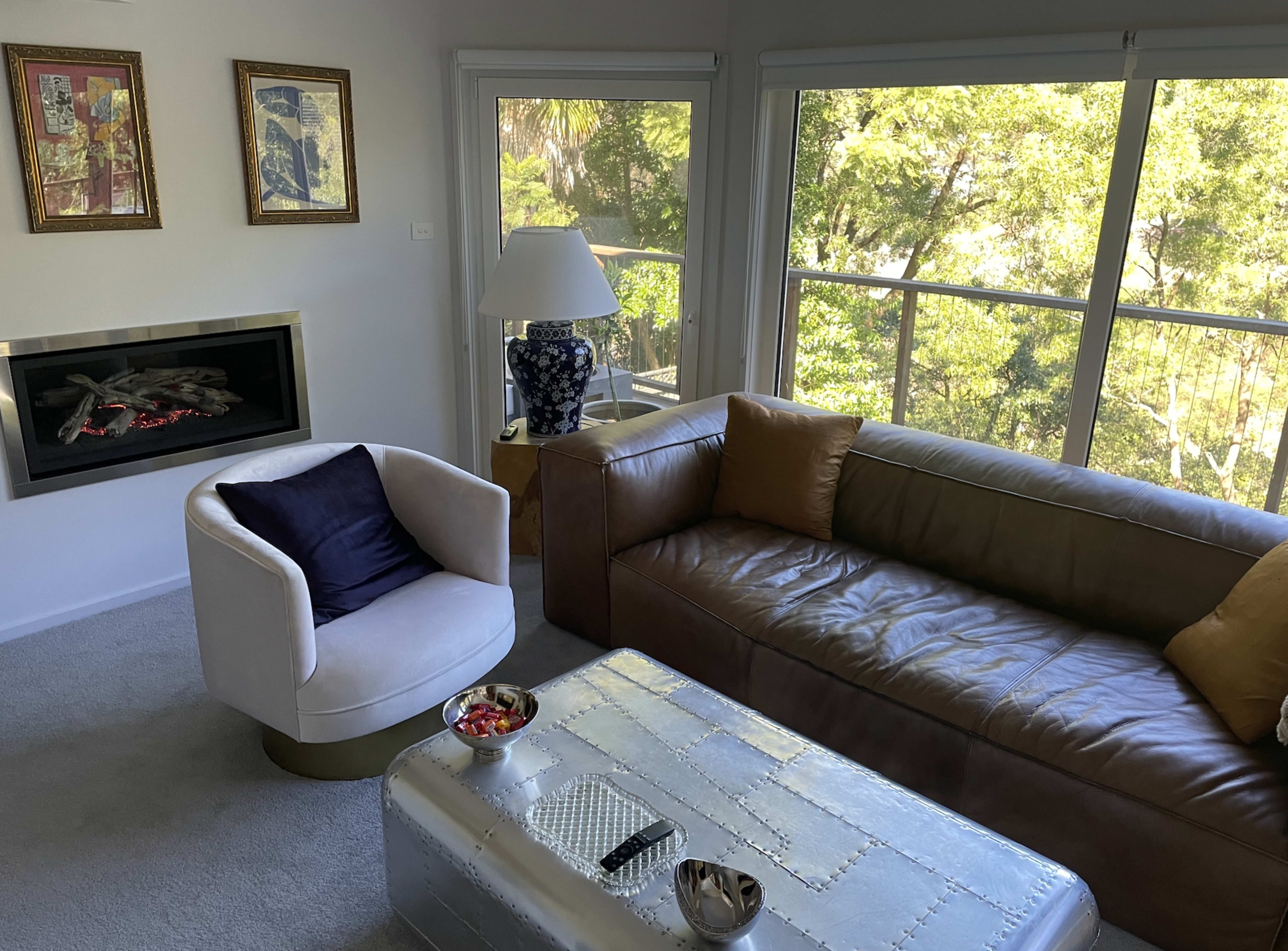A modern living room featuring a brown leather sofa, a white armchair, a silver coffee table, and a window with a view of greenery outside.