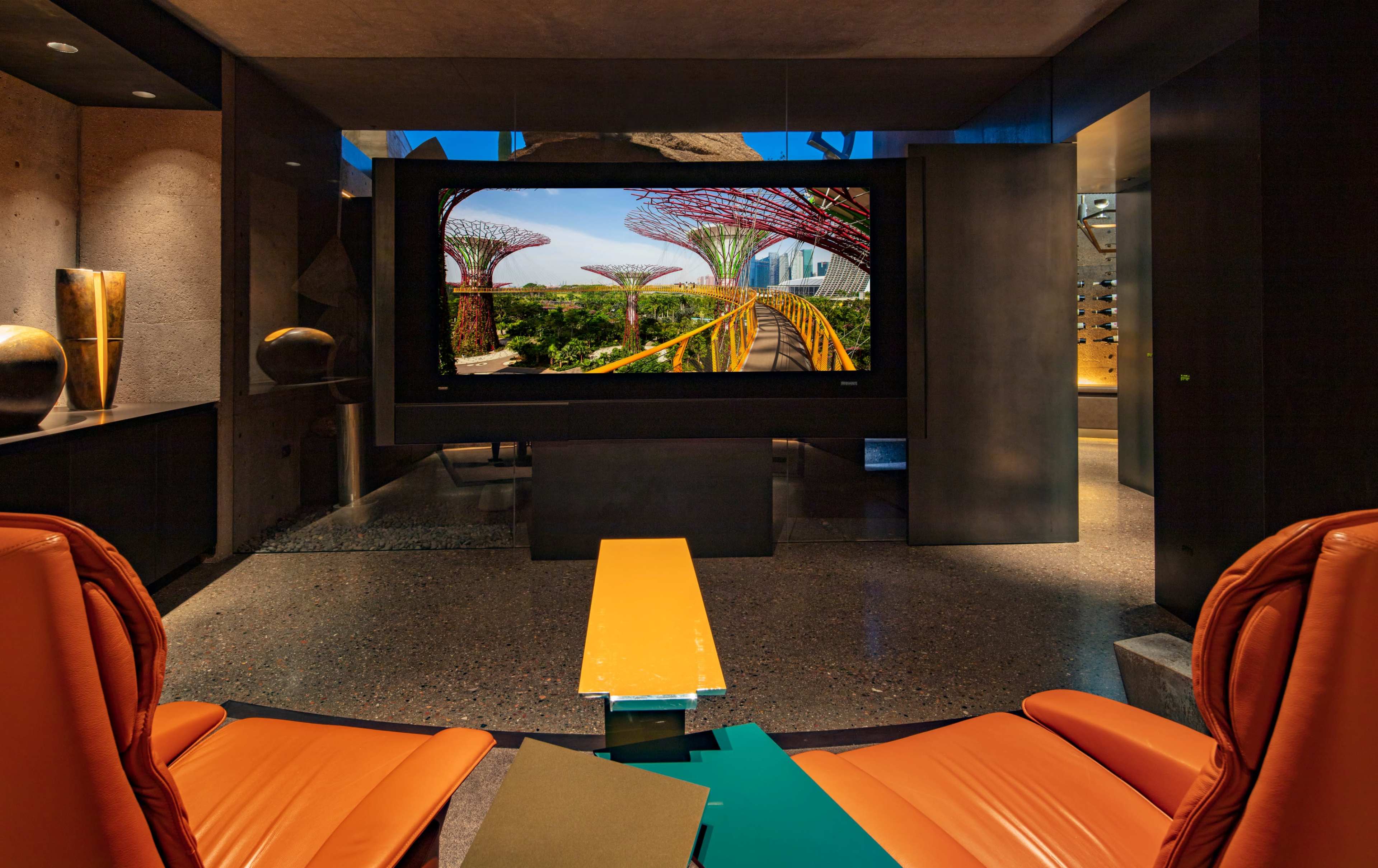 A modern interior featuring two orange chairs facing a large screen displaying a vibrant garden scene with tall structures in the background.
