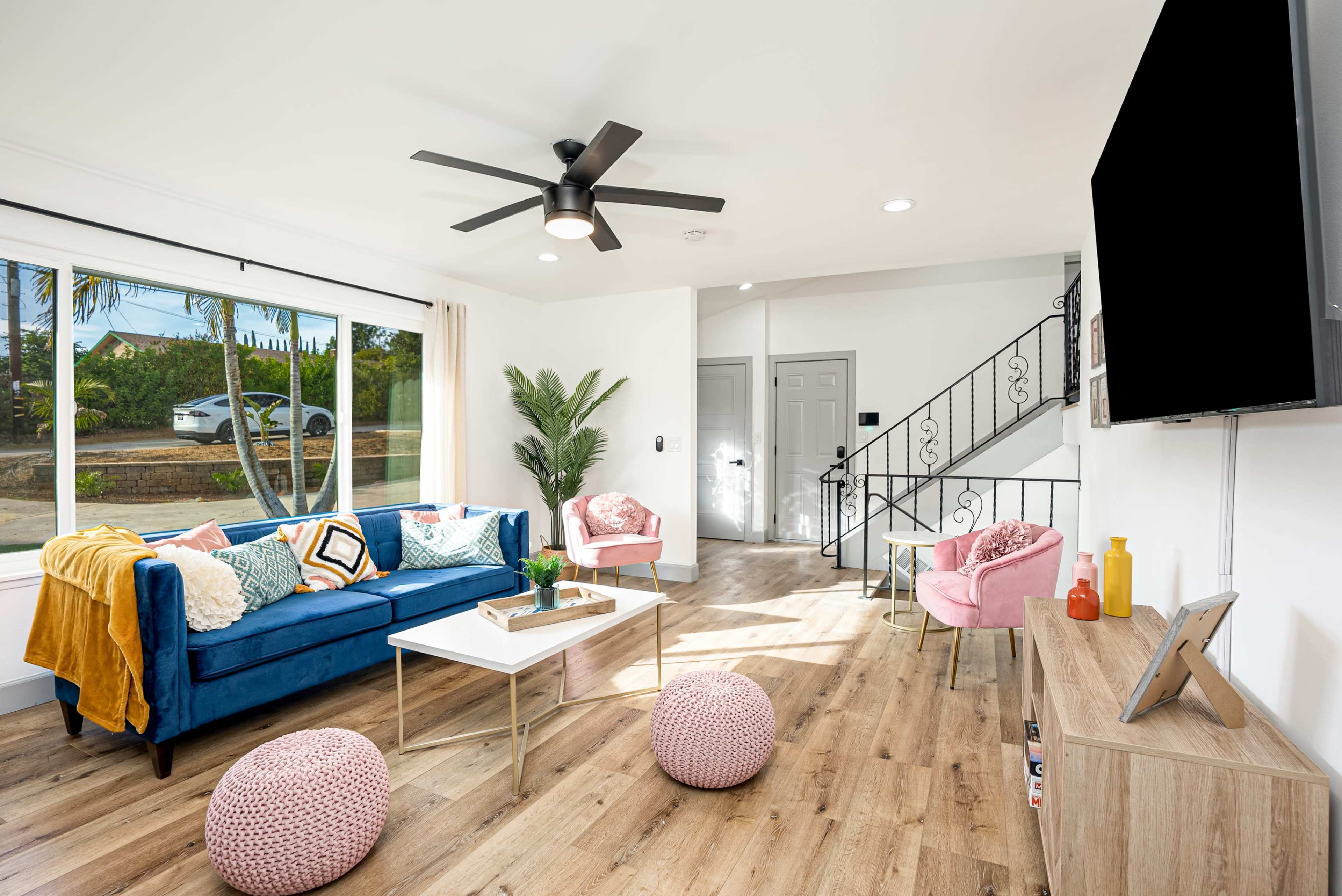 A bright living room features a blue sofa, two pink armchairs, a coffee table, and decorative poufs, with a staircase visible in the background and a large window offering outdoor views.