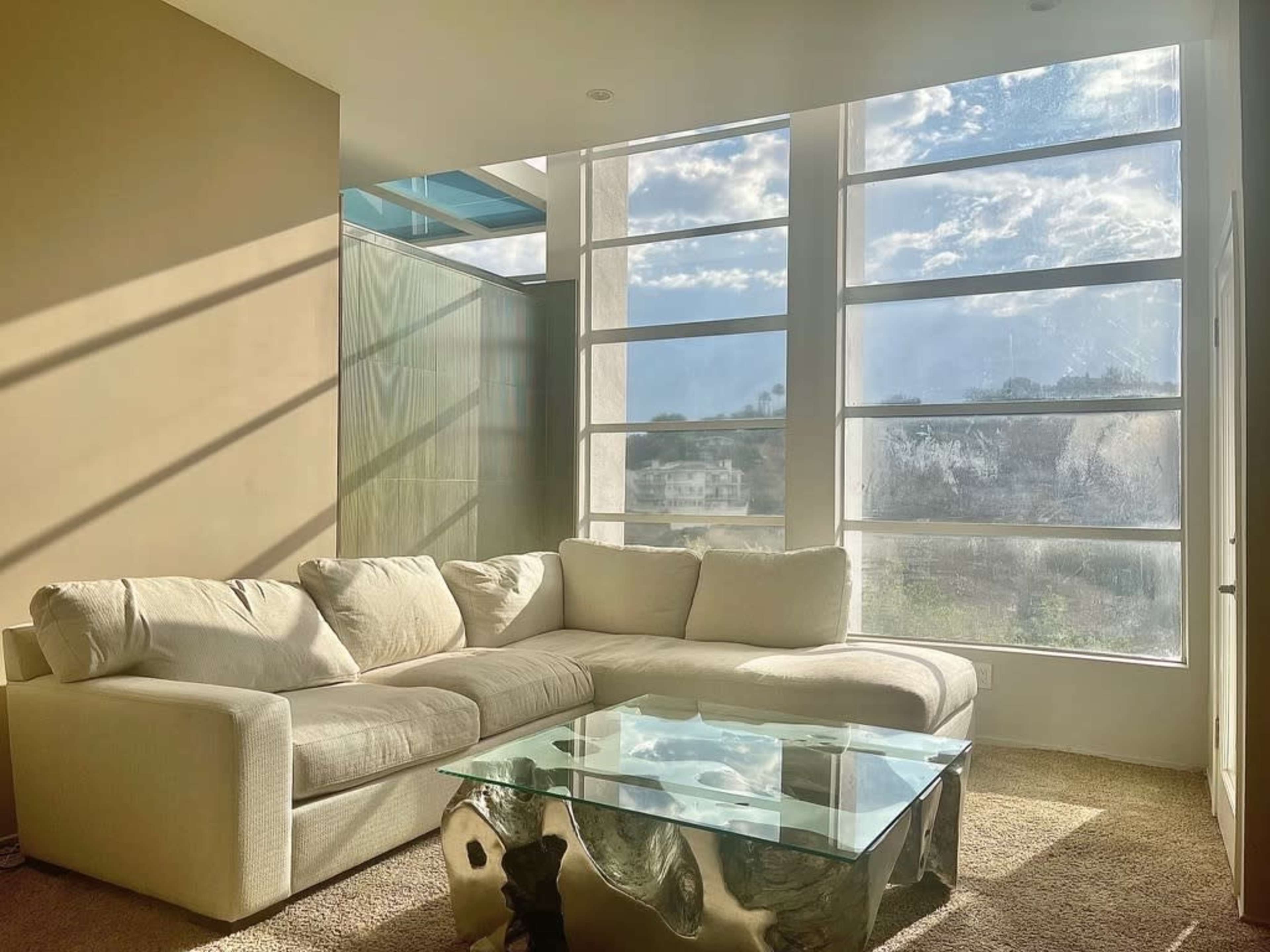 A modern living room features a white sectional sofa, a glass coffee table, and large windows allowing natural light to fill the space.