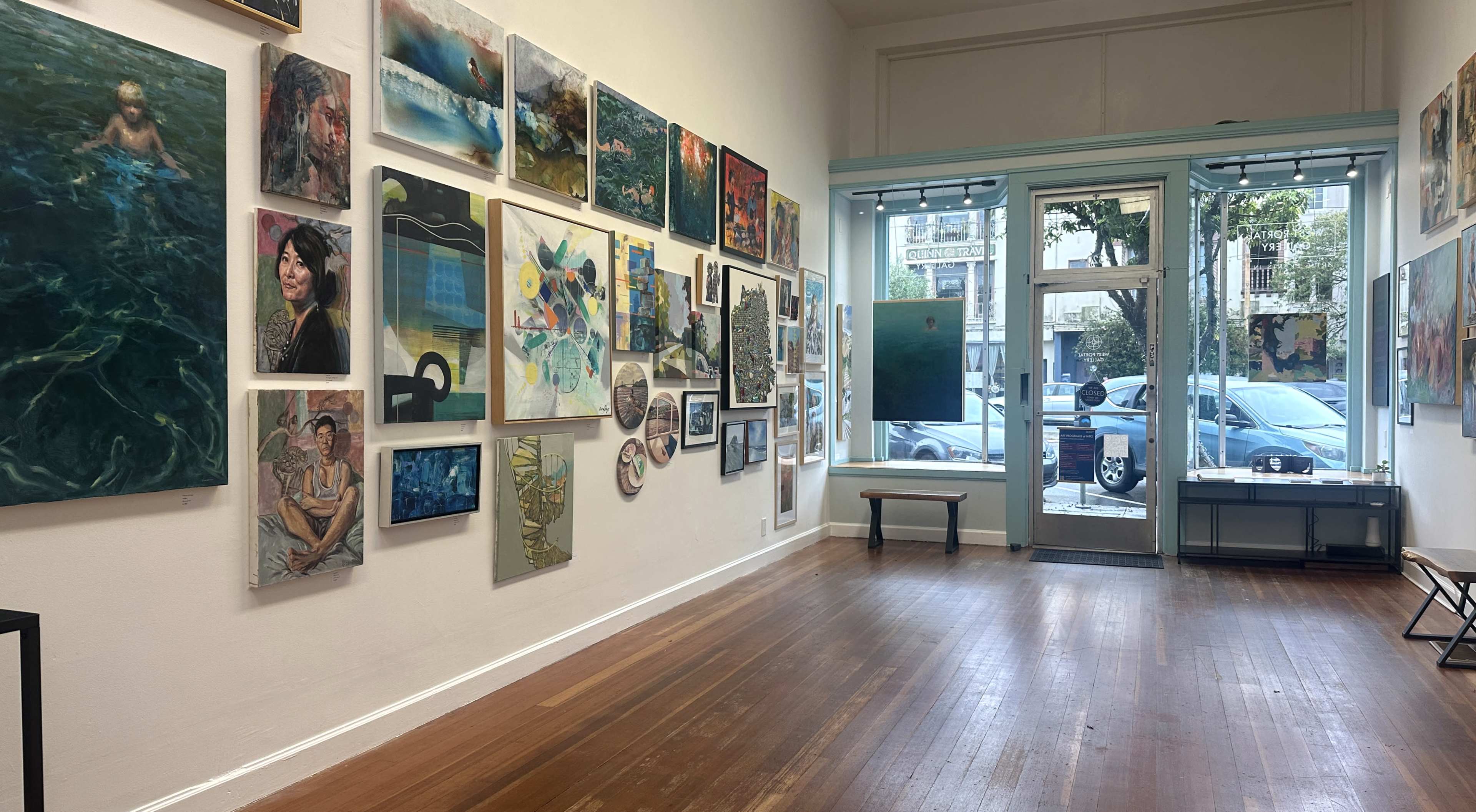 The image shows an art gallery with various paintings displayed on a white wall, and a large window at the front allowing natural light into the space.