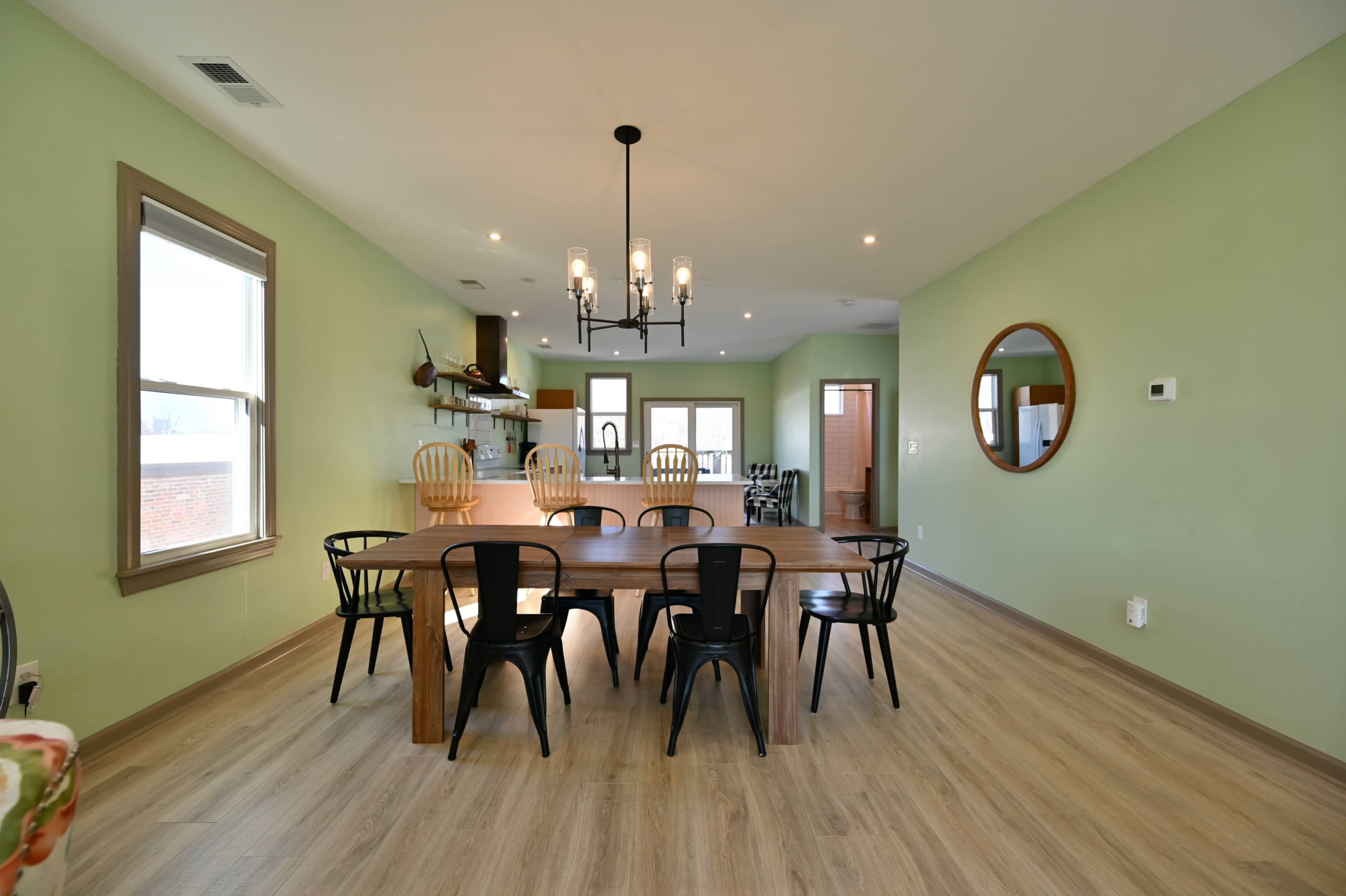 A spacious dining area features a long wooden table surrounded by black and wooden chairs, with green walls and modern lighting fixtures above.