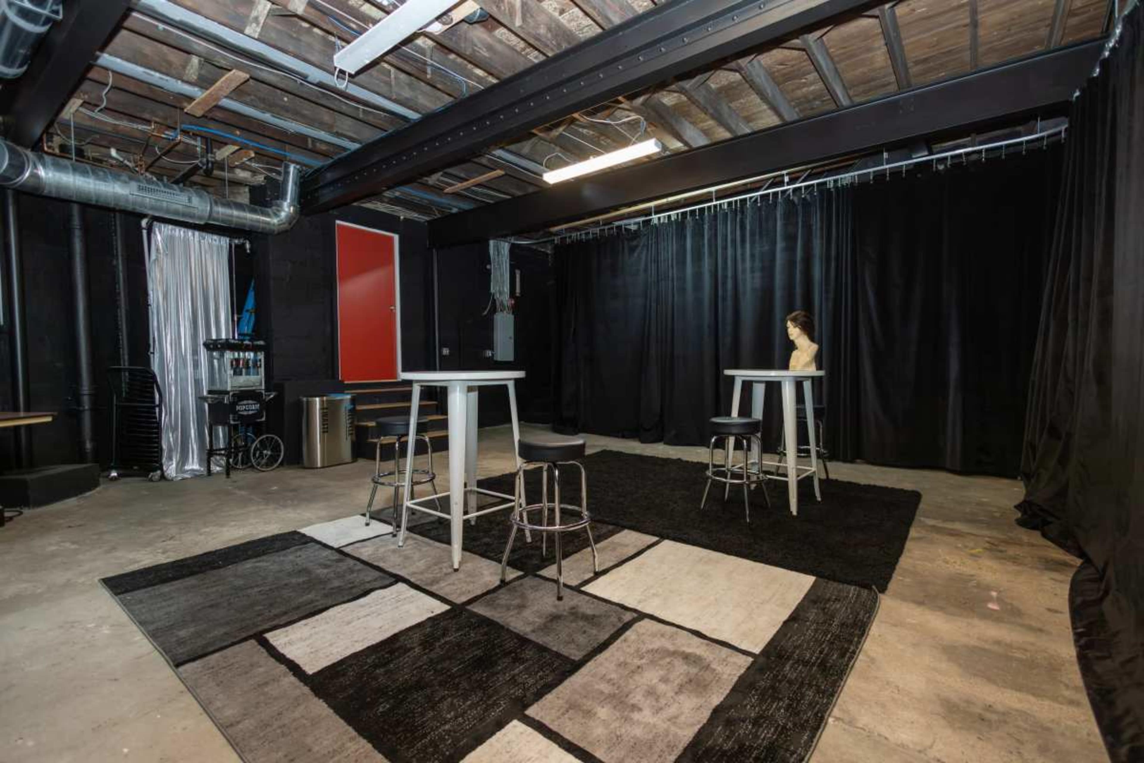 The image shows a dimly lit studio space with a concrete floor, featuring two tall tables surrounded by stools and a rug with a geometric design.