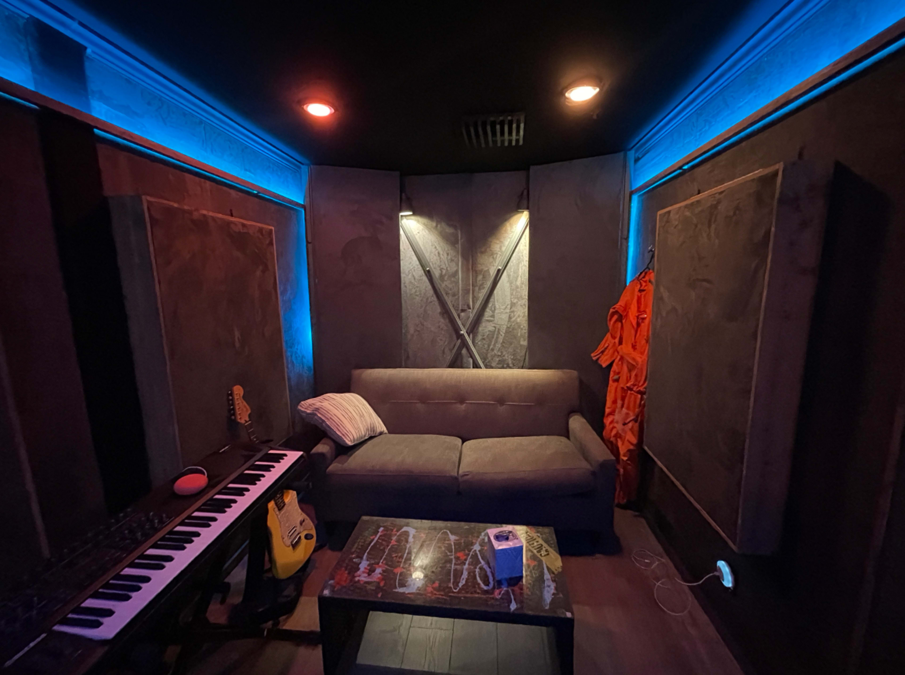 The interior of a music studio features a gray sofa, a keyboard, a guitar, and orange garments hanging on the wall, illuminated by blue and red lights.