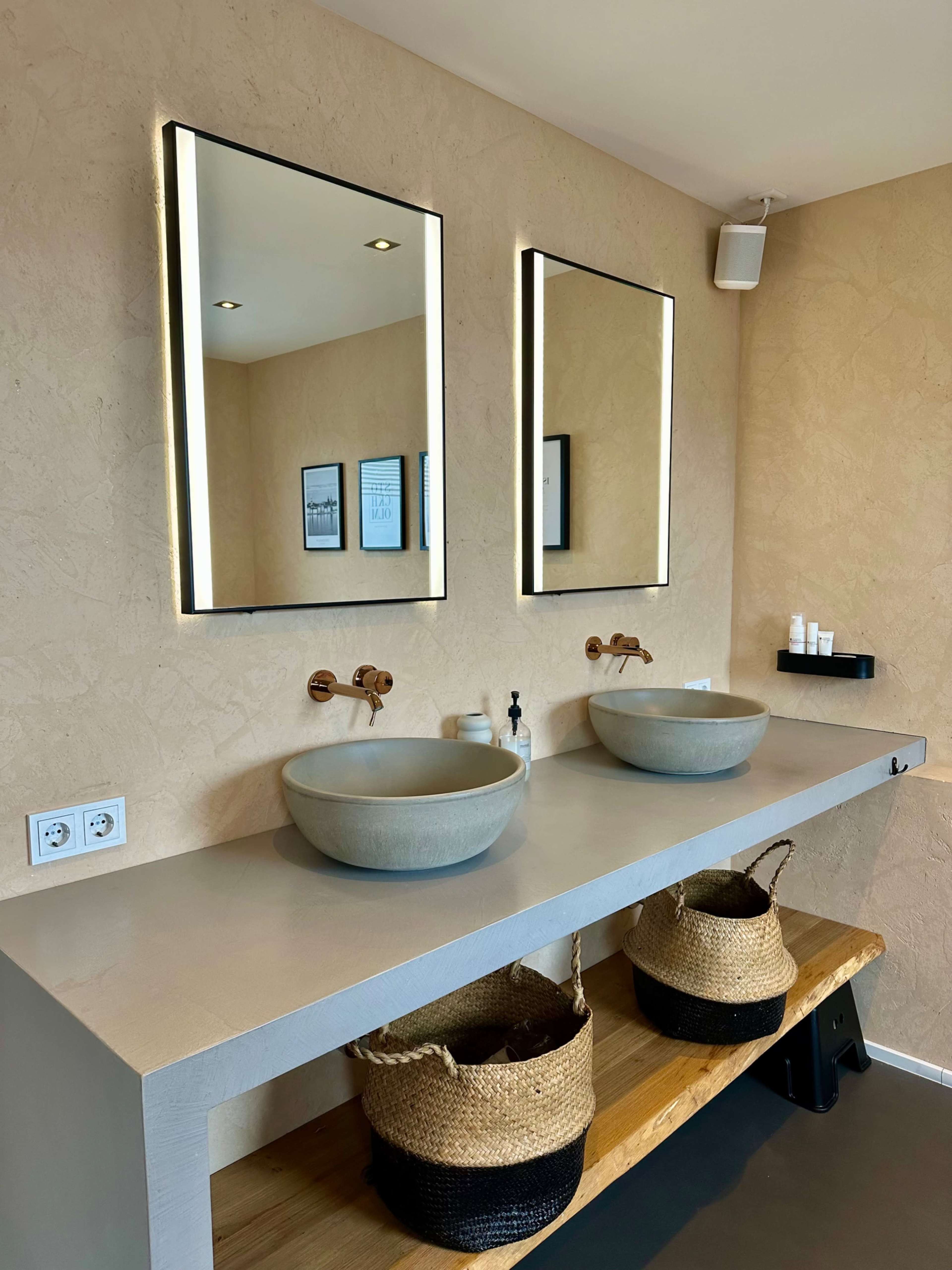 The image shows a modern bathroom with two round bowl sinks, backlit mirrors, and woven baskets underneath a wooden shelf.